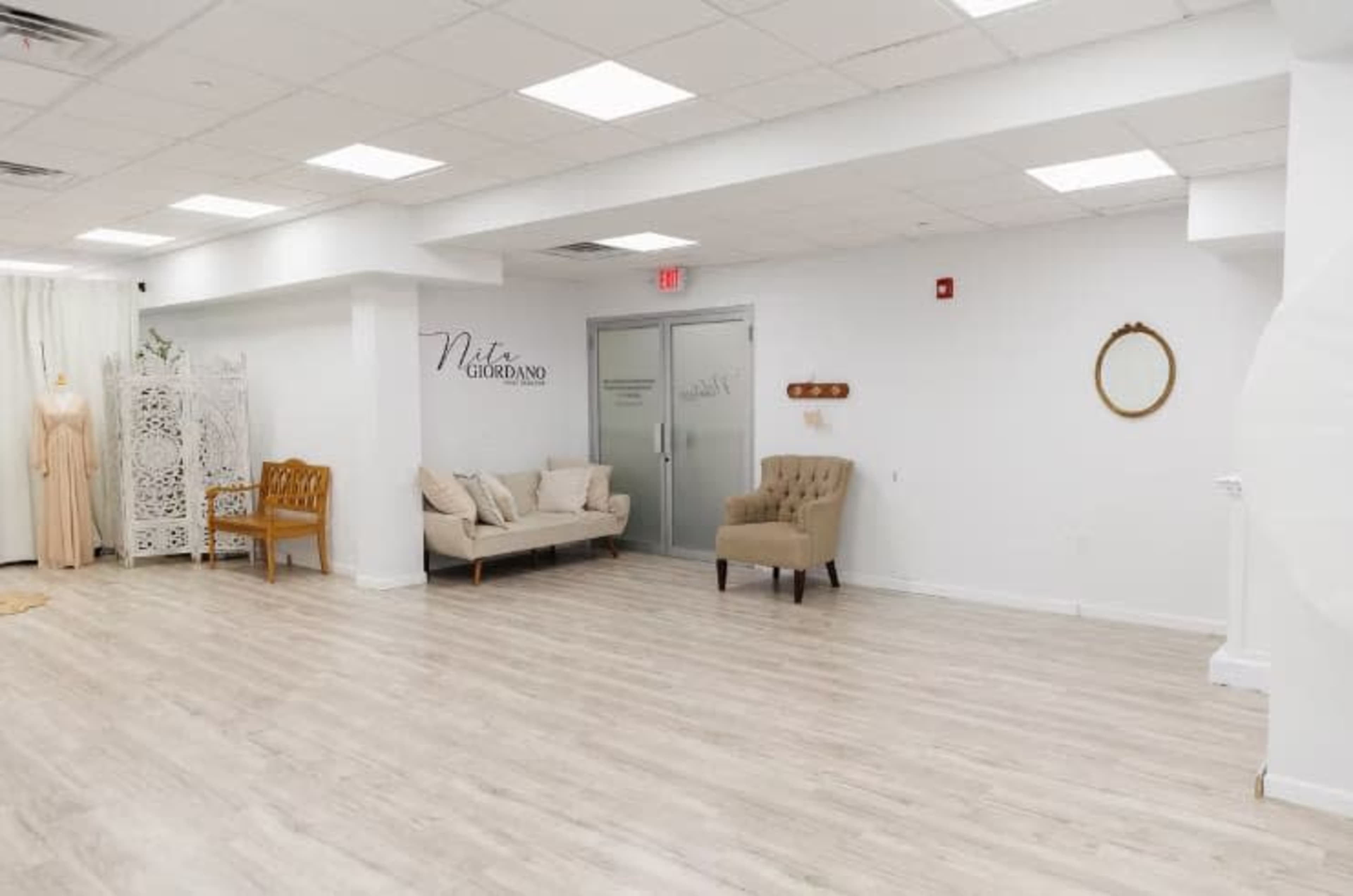 The image shows a spacious, well-lit room with light wooden floors, featuring two pieces of seating furniture, a full-length mirror, and minimal decorative elements on the walls.
