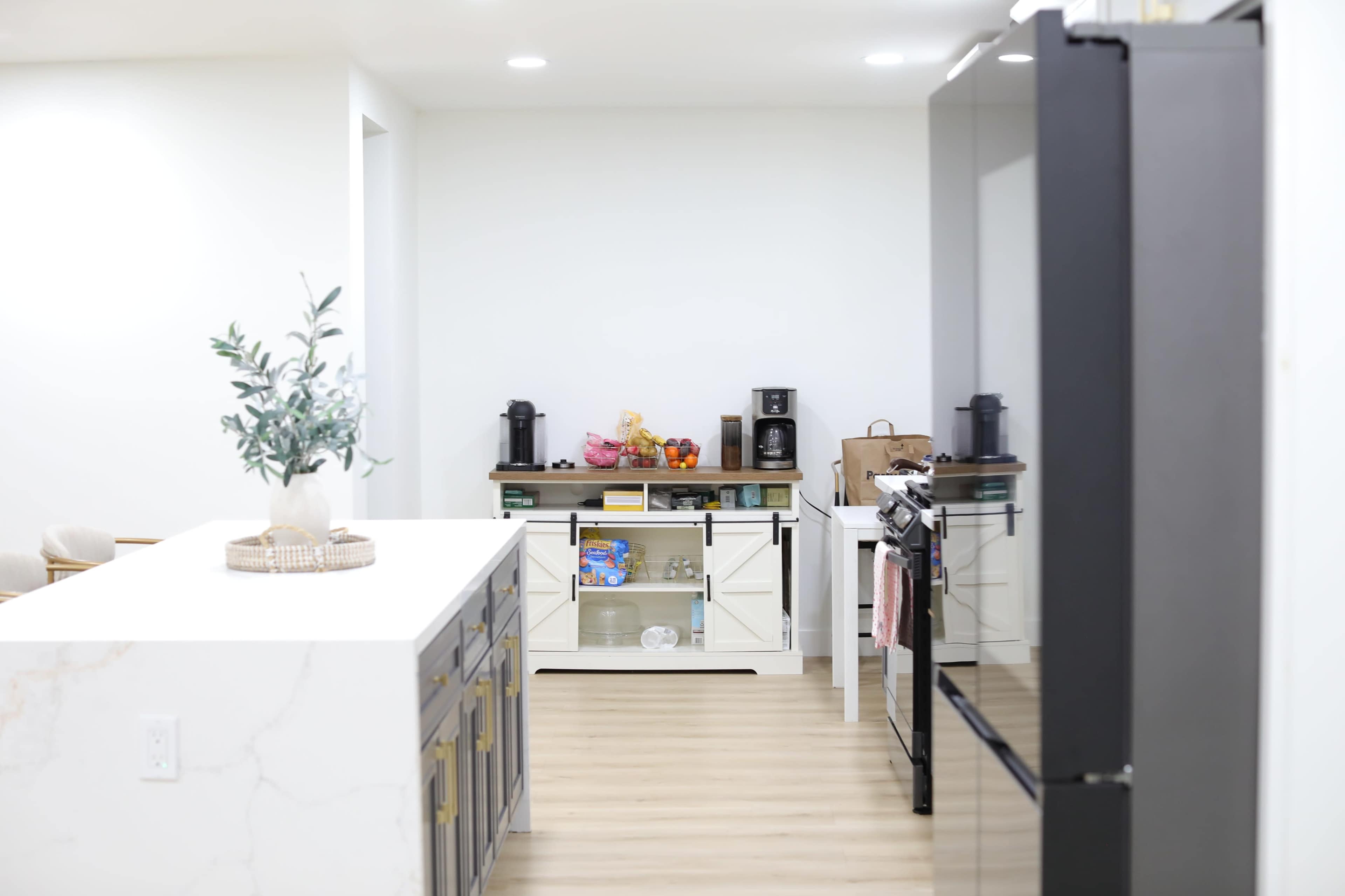 The image shows a modern kitchen featuring a dark refrigerator, a white countertop island, and an organized shelf displaying kitchen appliances and snacks.