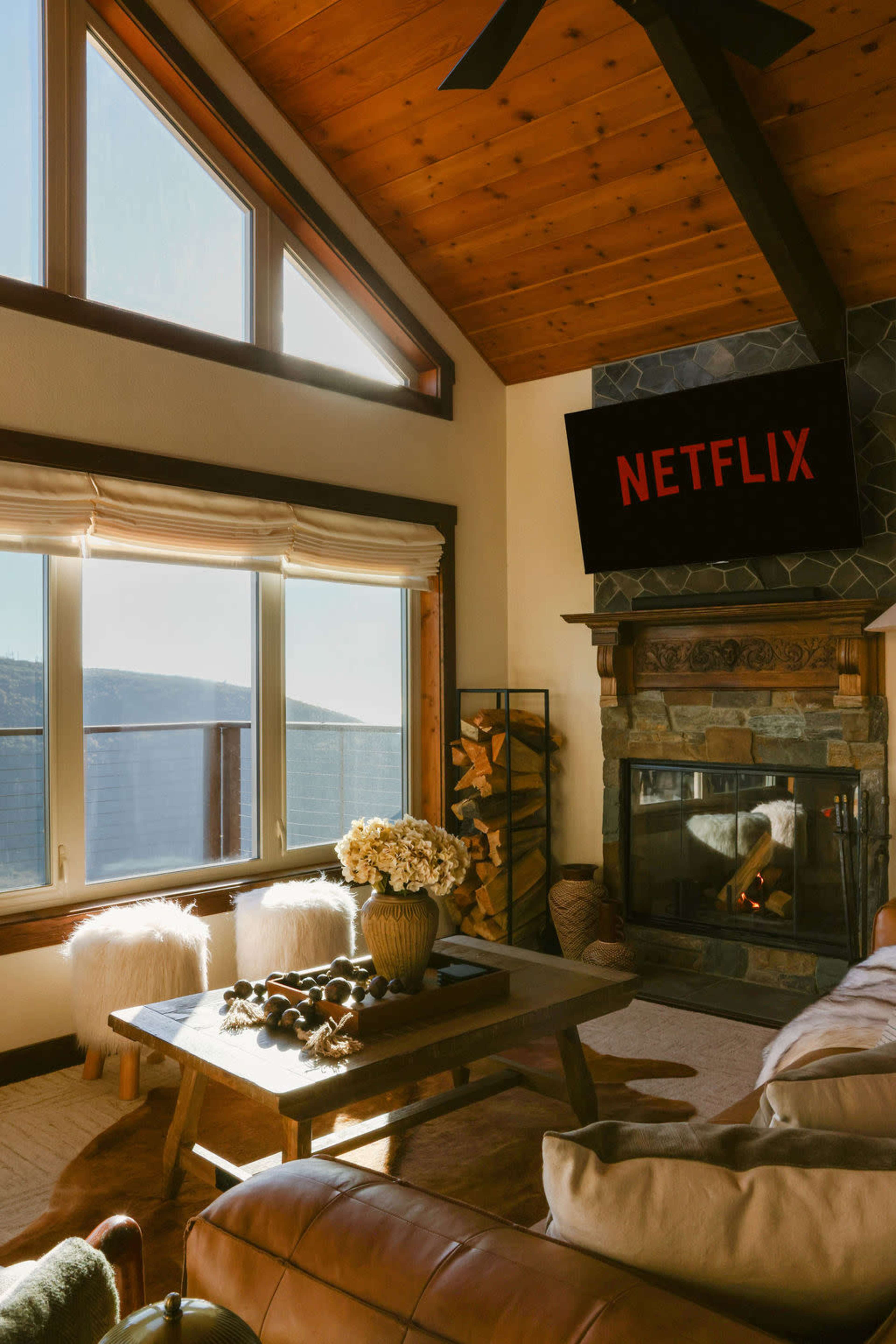 The image shows a cozy living room with large windows, a stone fireplace, a wooden coffee table adorned with decorative items, and a wall-mounted TV displaying the Netflix logo.