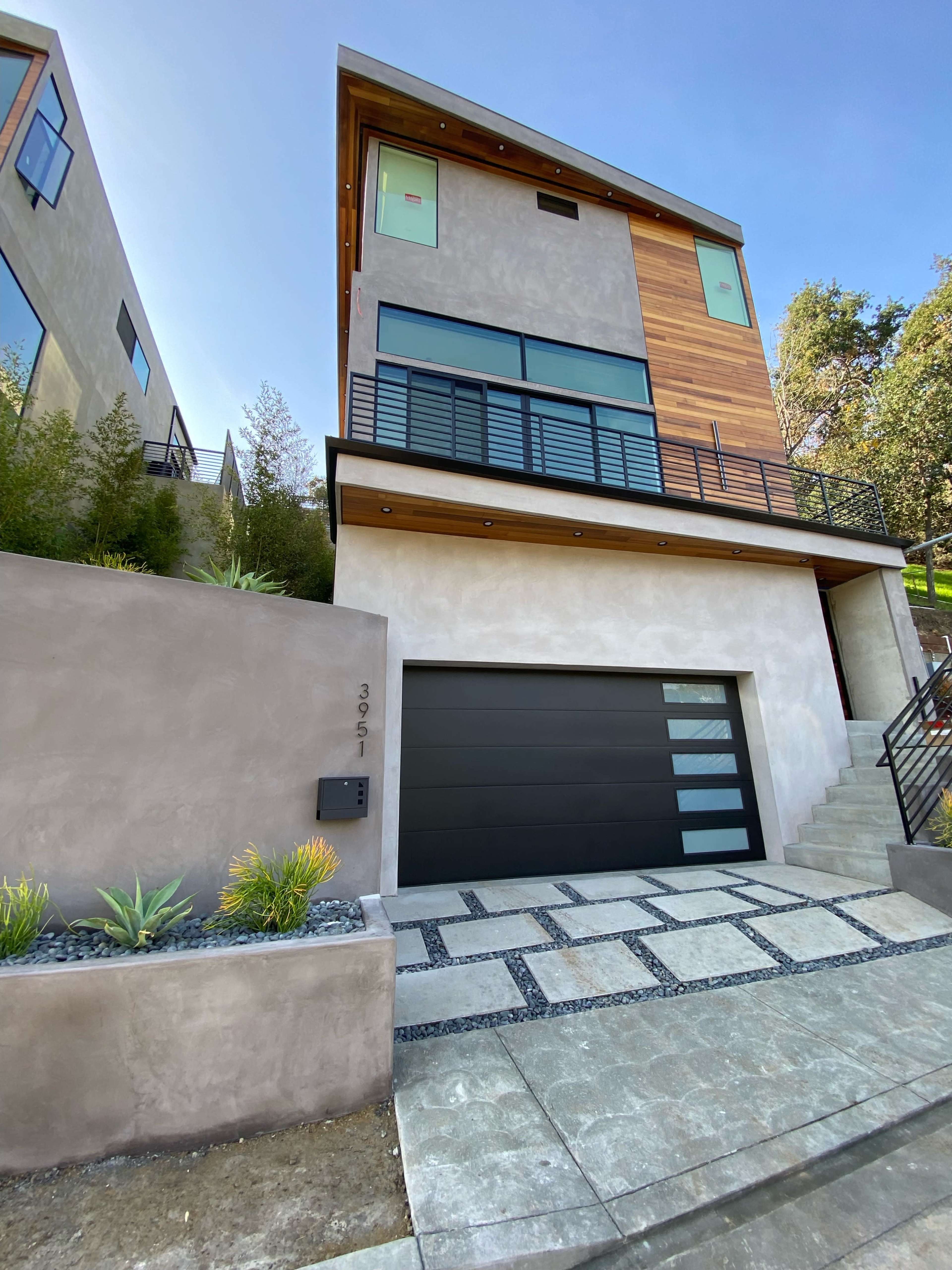 A modern three-story house with a wood-accented upper section and a gray stucco facade features a large garage door and a stone pathway leading to the entrance.