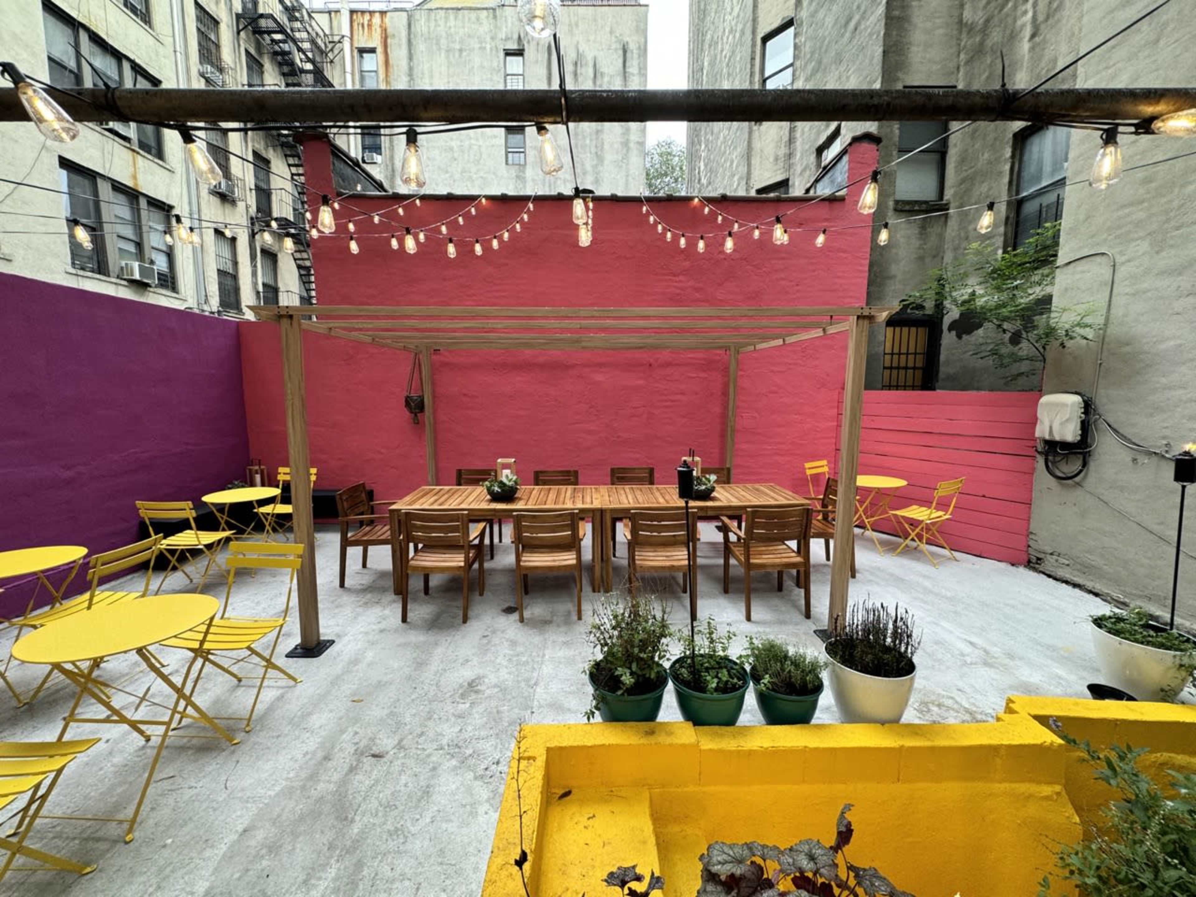 An outdoor patio space featuring yellow metal chairs, wooden tables, and a pink wall, illuminated by string lights.