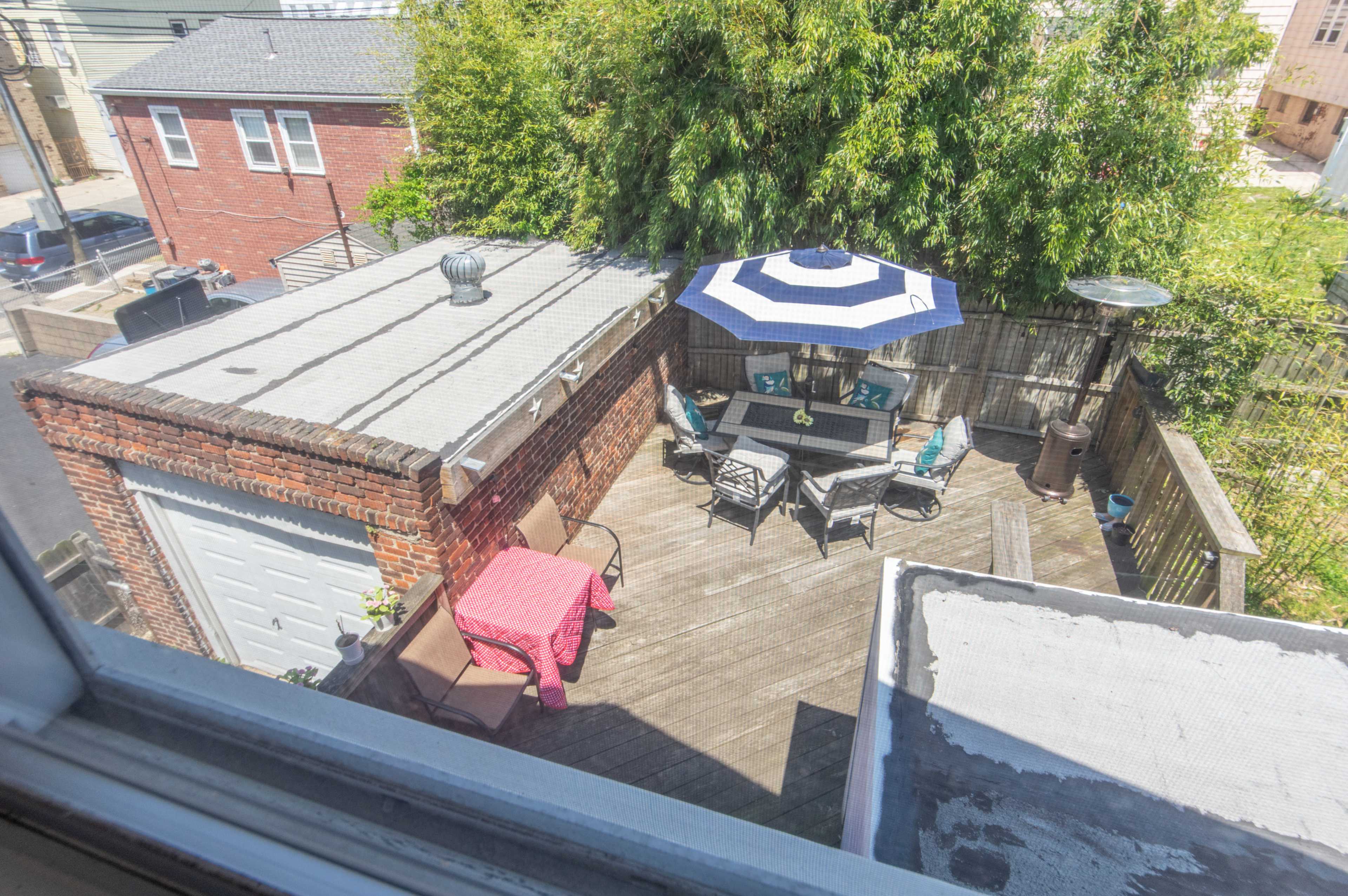 A rooftop patio with a table and chairs, featuring a large umbrella, surrounded by greenery and adjacent buildings.