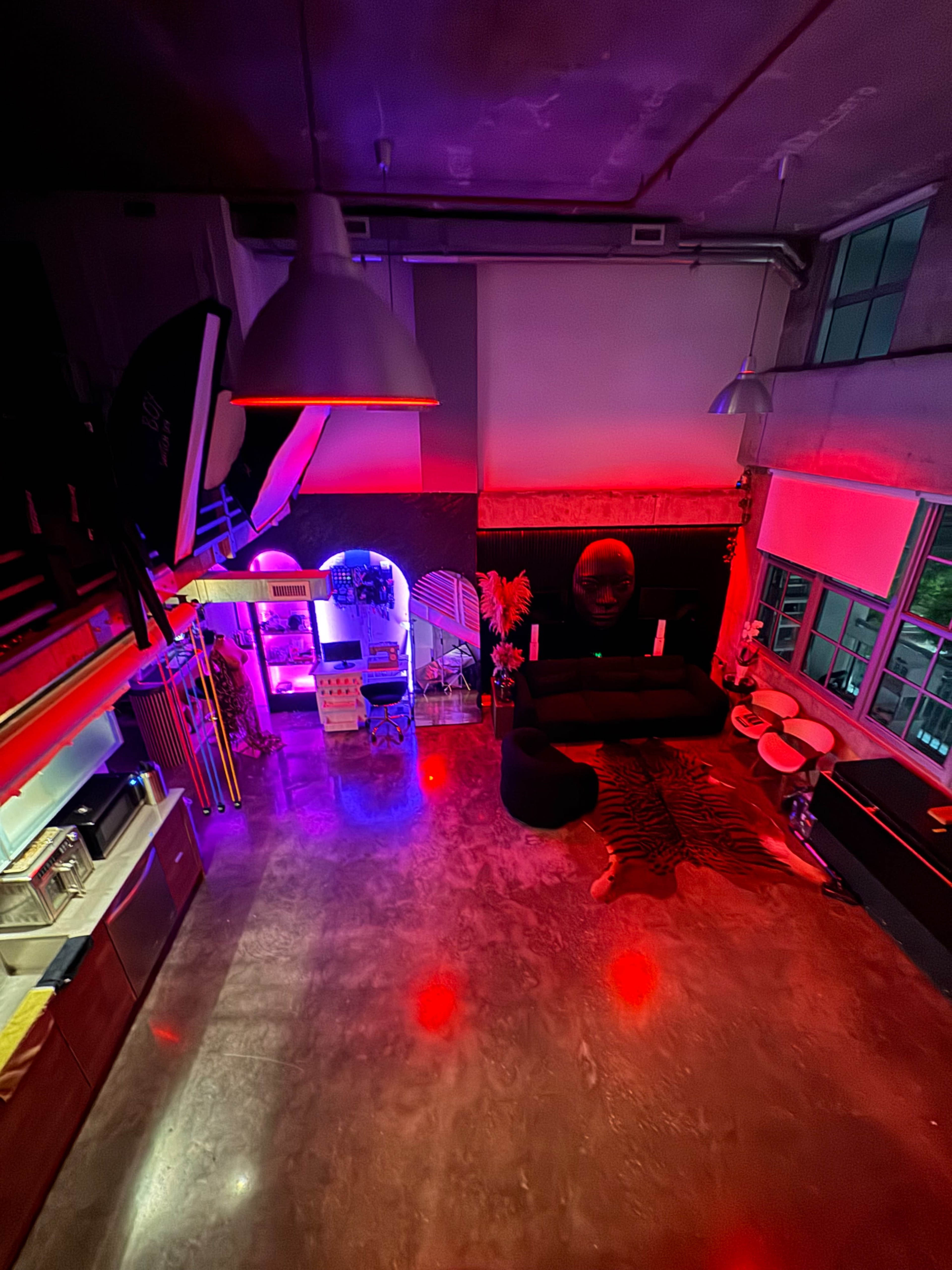 The image shows a modern, industrial-style loft with various areas illuminated by red and purple lights, featuring a kitchen, seating area, and decorative elements.