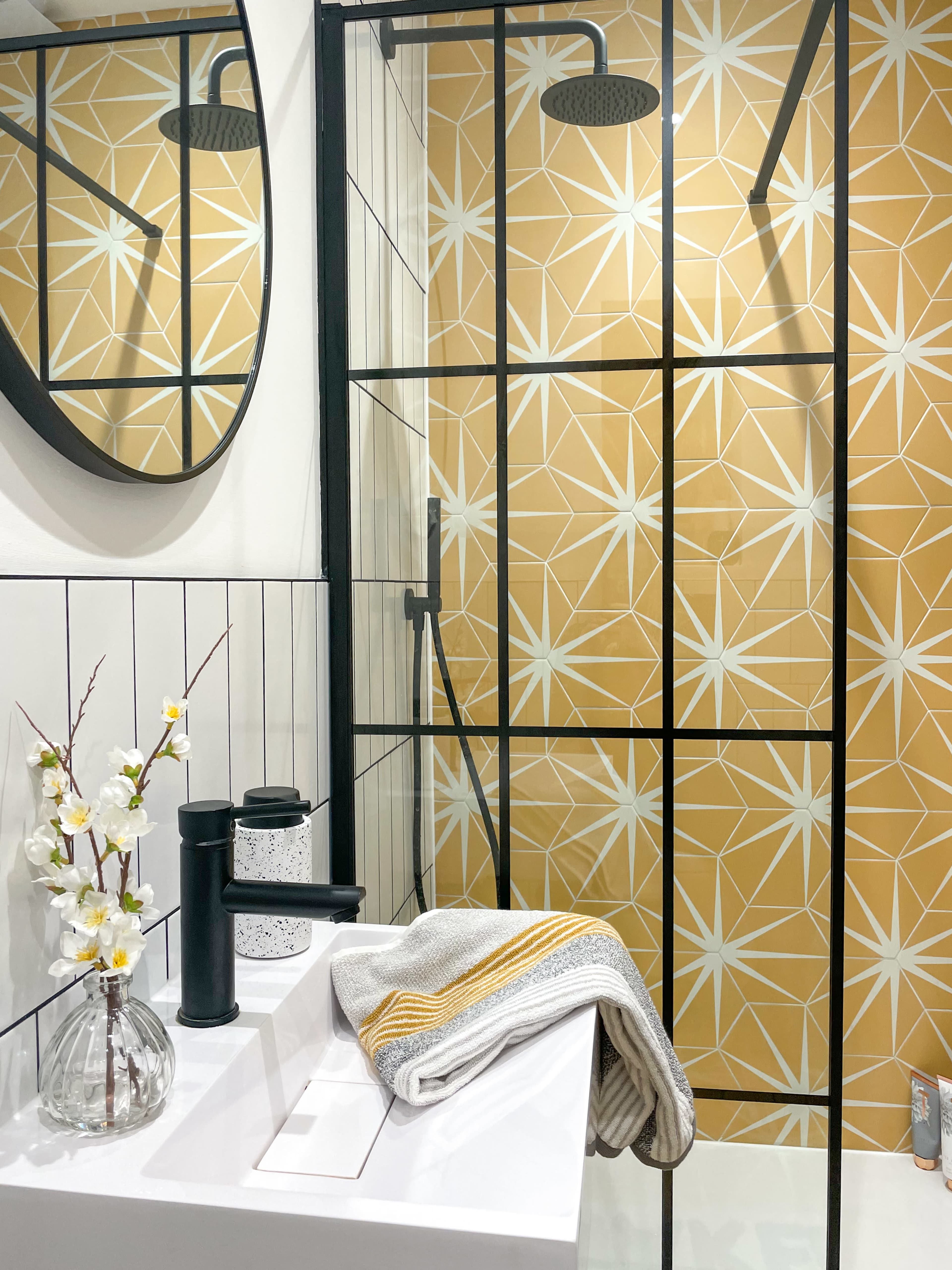 The image shows a modern shower space with a yellow geometric patterned wall, a round mirror, a sleek faucet, and a vase with flowers on a countertop.