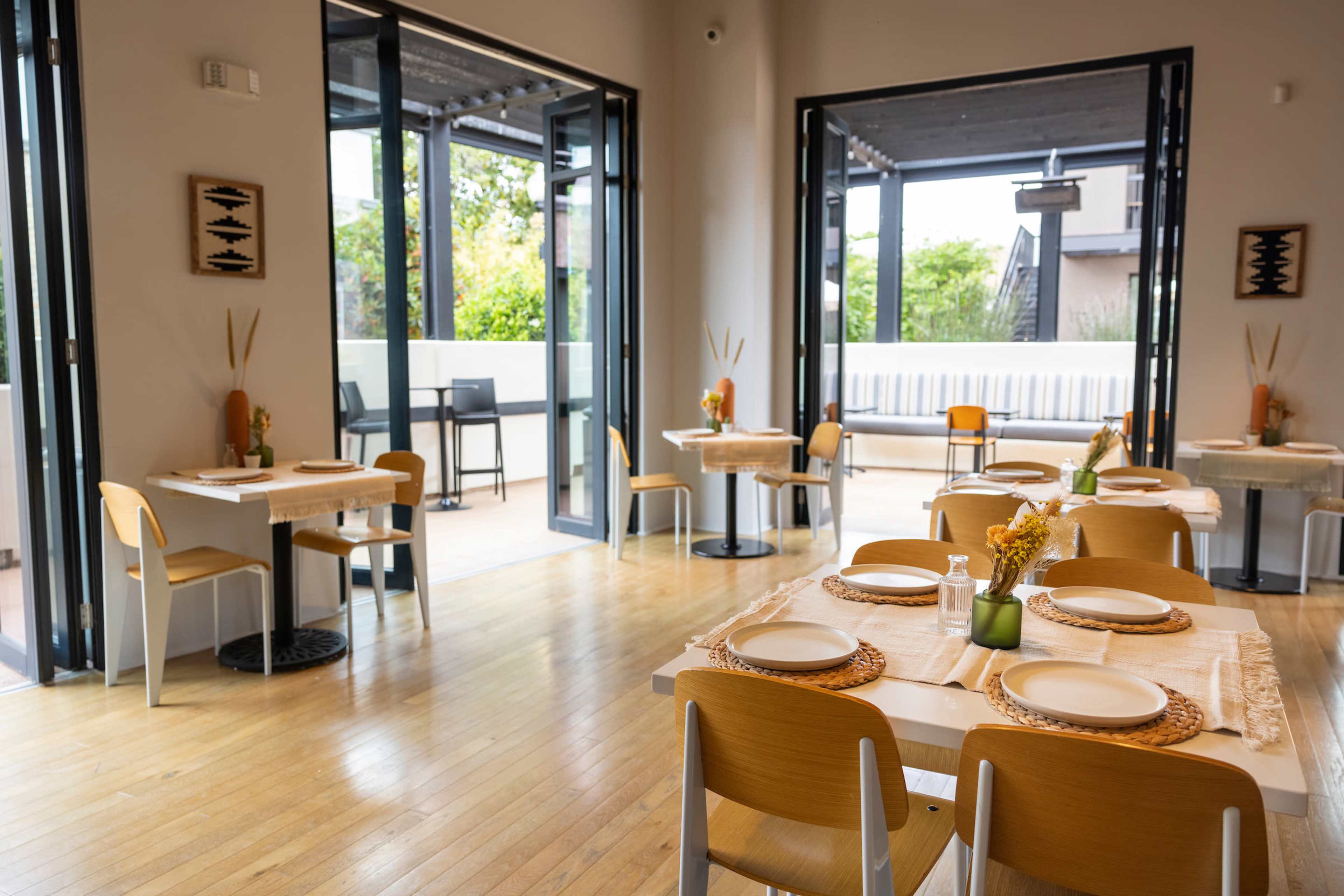 A contemporary restaurant interior features several tables set for dining, with floor-to-ceiling windows allowing natural light to fill the space.