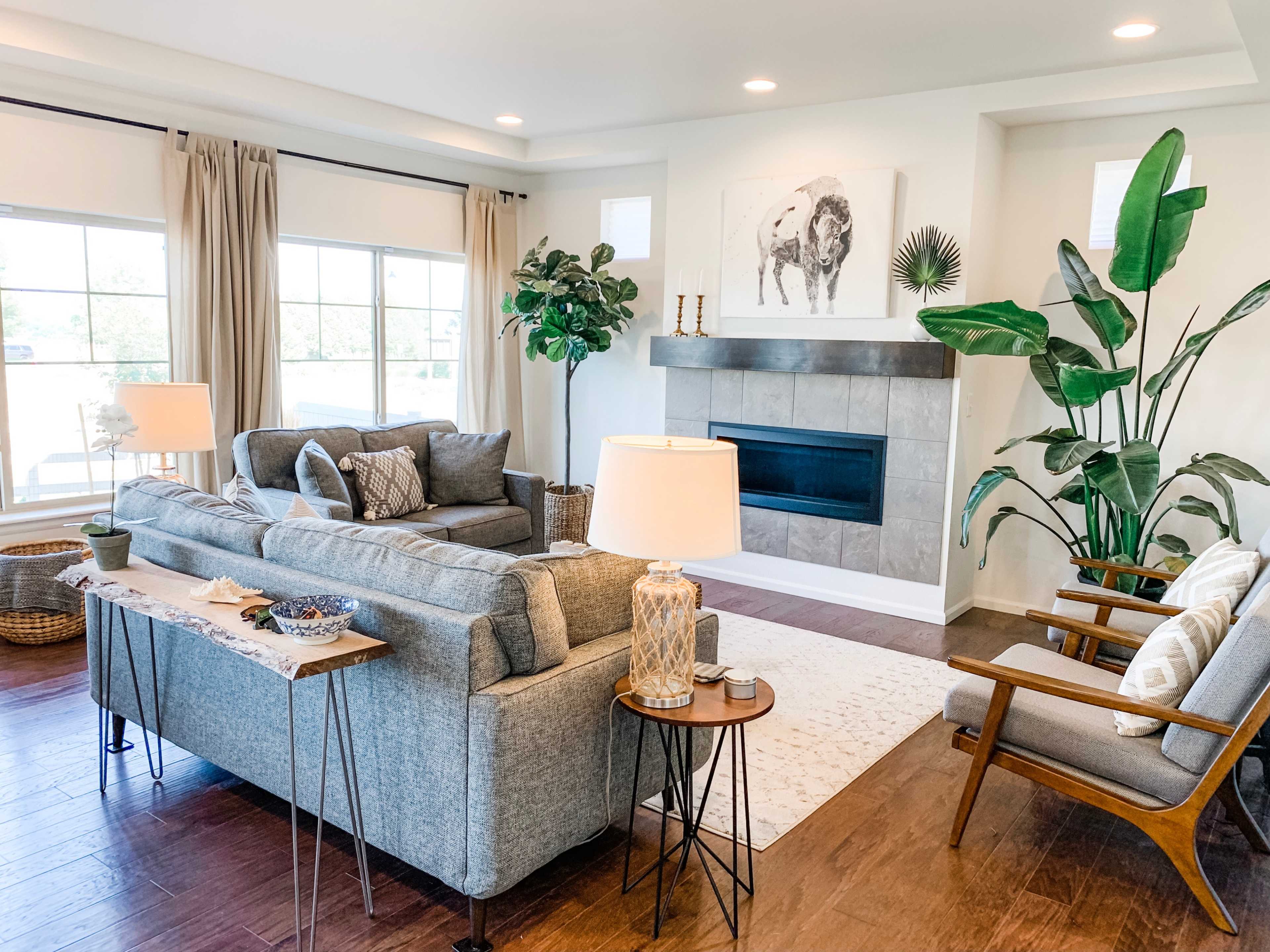 A cozy living room features a gray sofa, wooden chairs, a fireplace, and large windows with natural light.