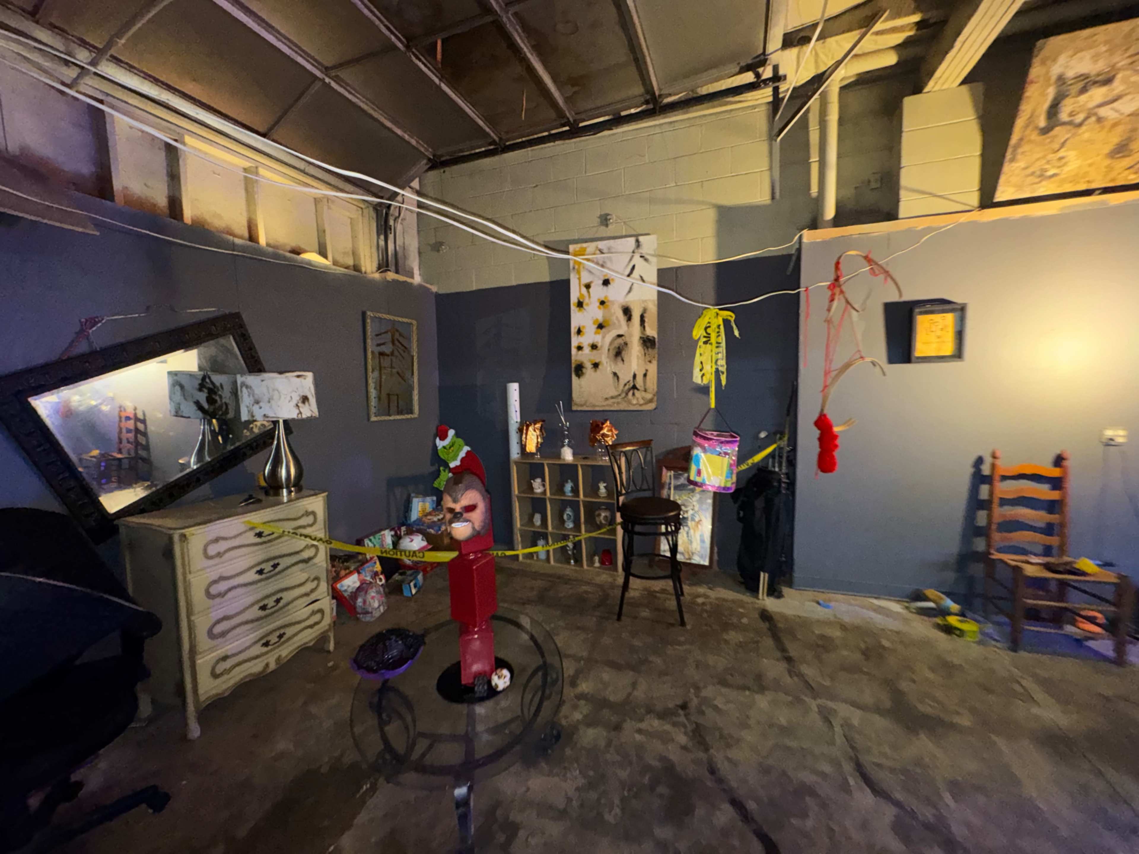 The image shows a dimly lit room with gray walls, featuring a mix of furniture, a large mirror, artwork on the walls, and various objects scattered on the floor.