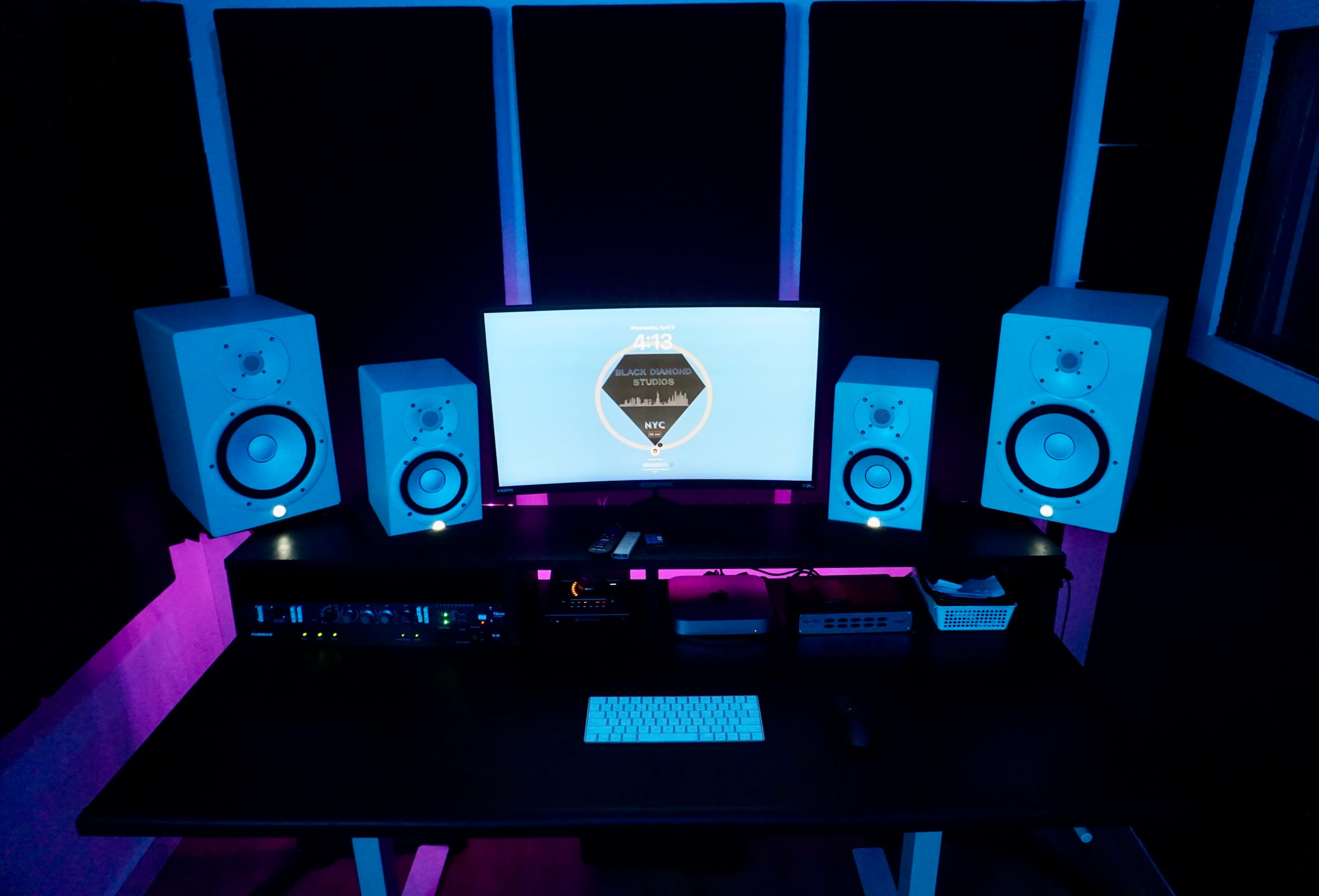 The image shows a professional music studio setup with two speakers on either side of a computer monitor, a sound interface on the desk, and soundproofing panels on the walls.