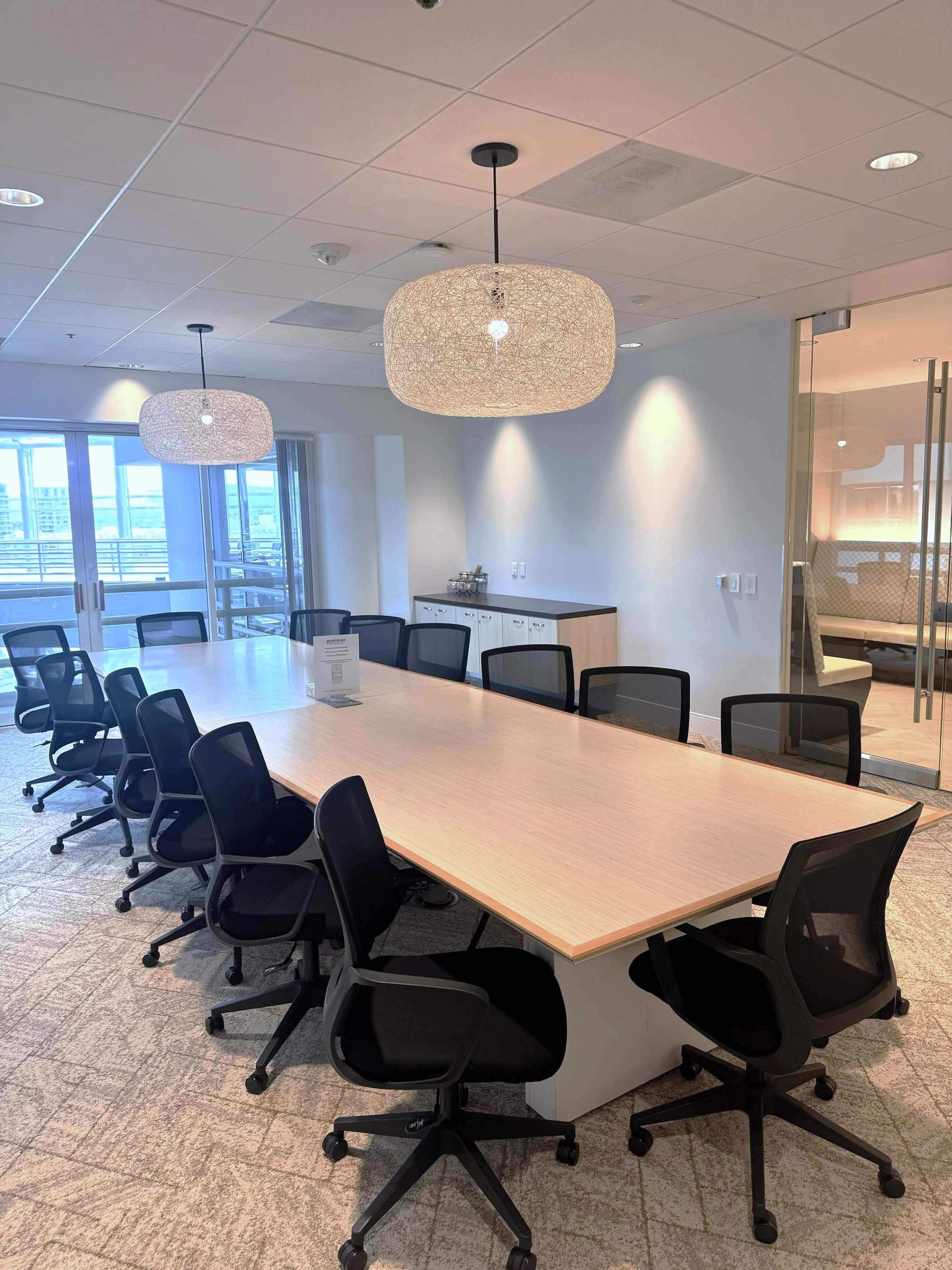 The image shows a modern conference room with a long wooden table surrounded by black ergonomic chairs and two decorative light fixtures hanging from the ceiling.