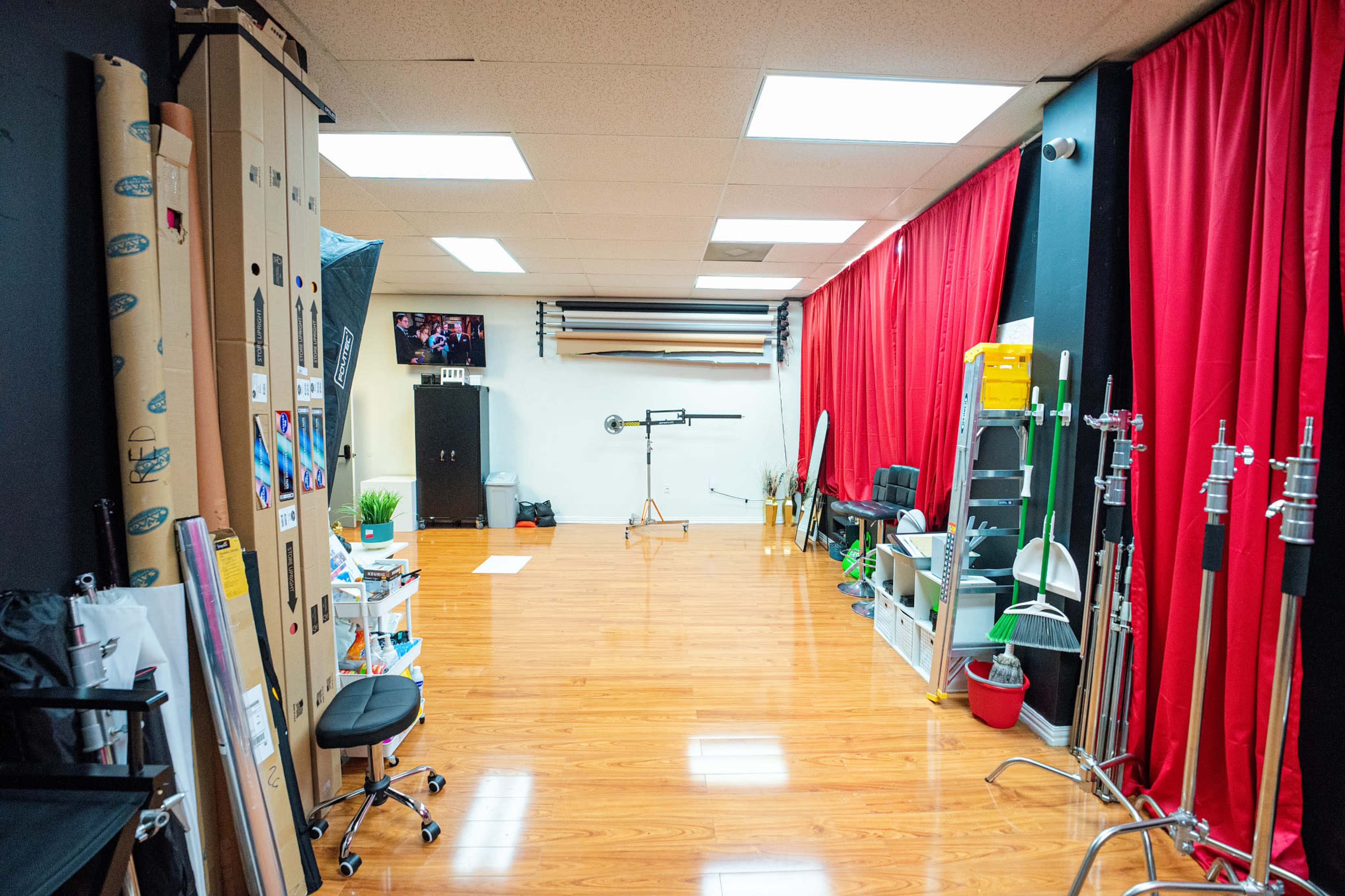 The image shows a spacious studio with wooden flooring, red curtains on one side, and various photography equipment and props neatly arranged along the walls.