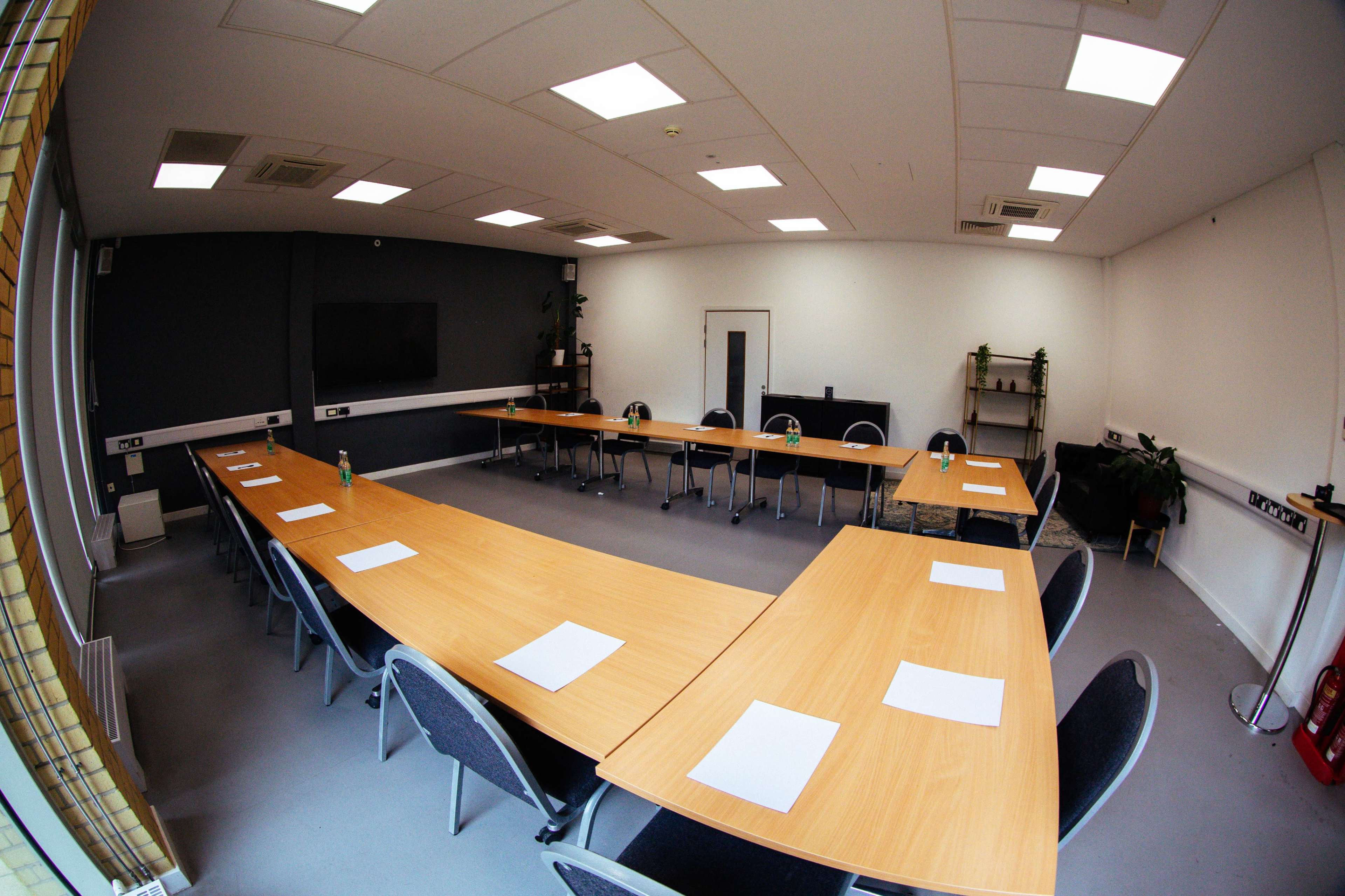 A large, empty conference room features multiple long wooden tables arranged in a U-shape with chairs around them and a screen mounted on the wall.