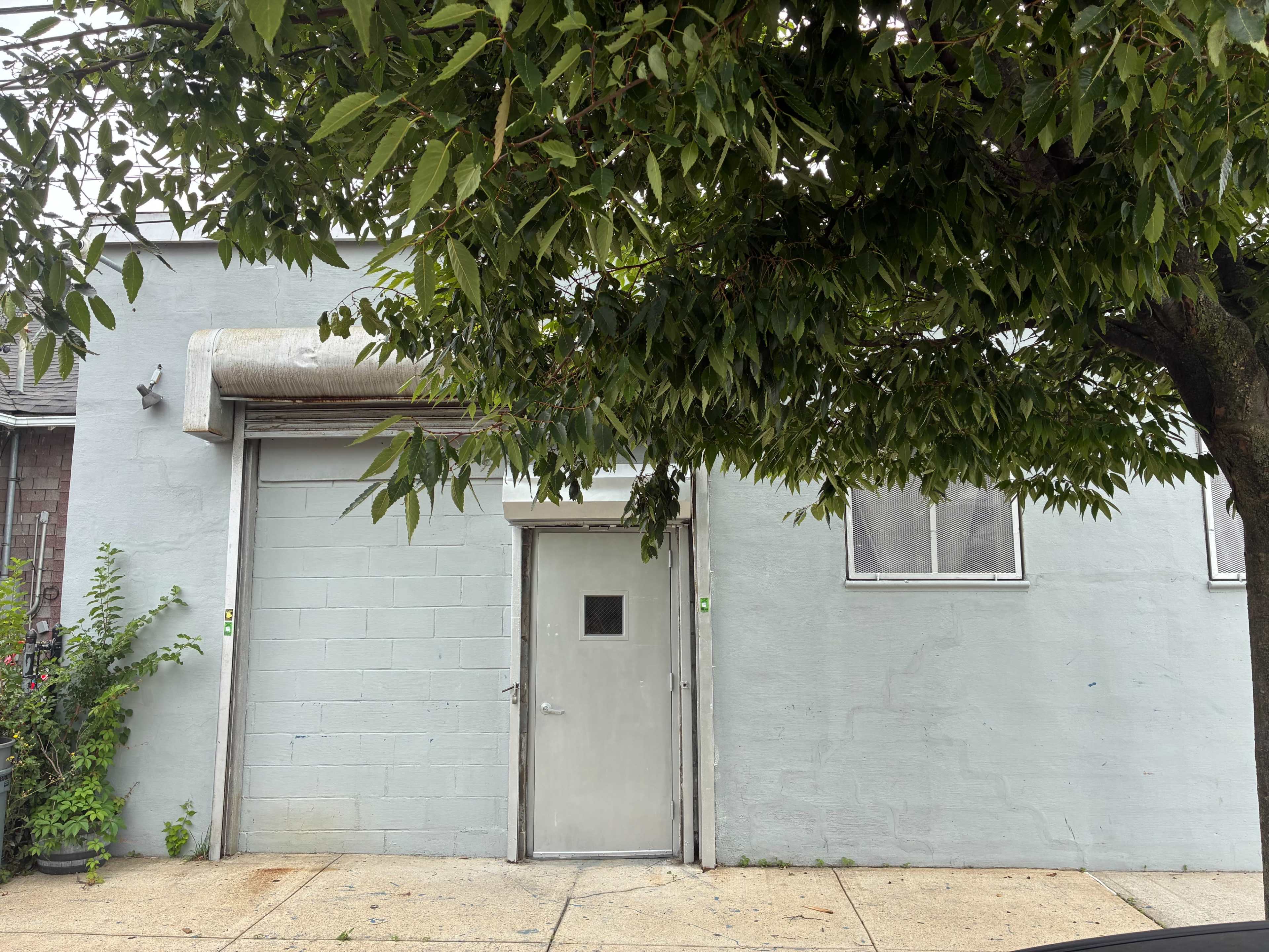 The image shows a pale gray building with a metal door and an overhead rolling door, partially obscured by a tree.