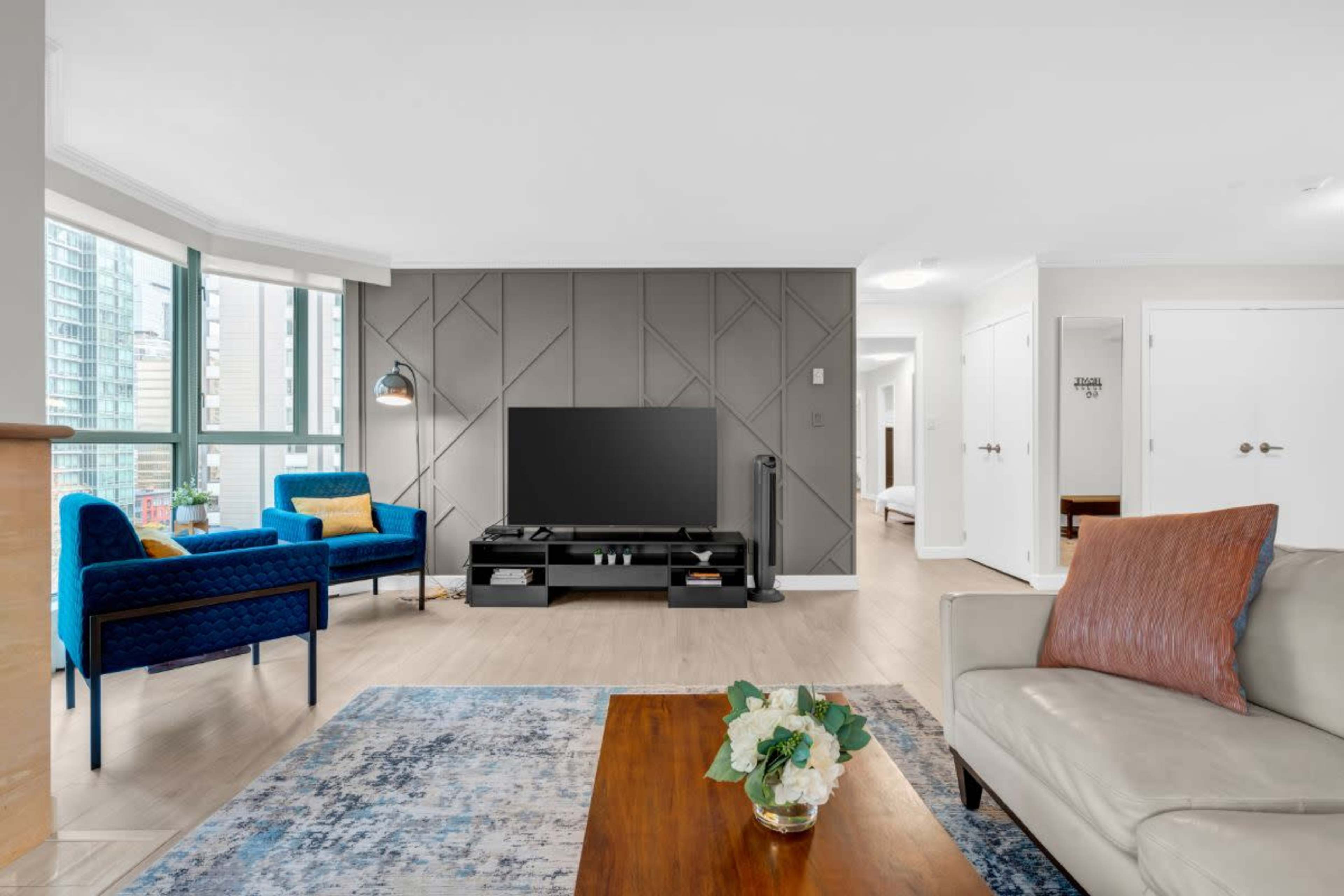 The image shows a modern living room featuring a large television, a light-colored sofa, blue armchairs, and a decorative coffee table with a vase of flowers.