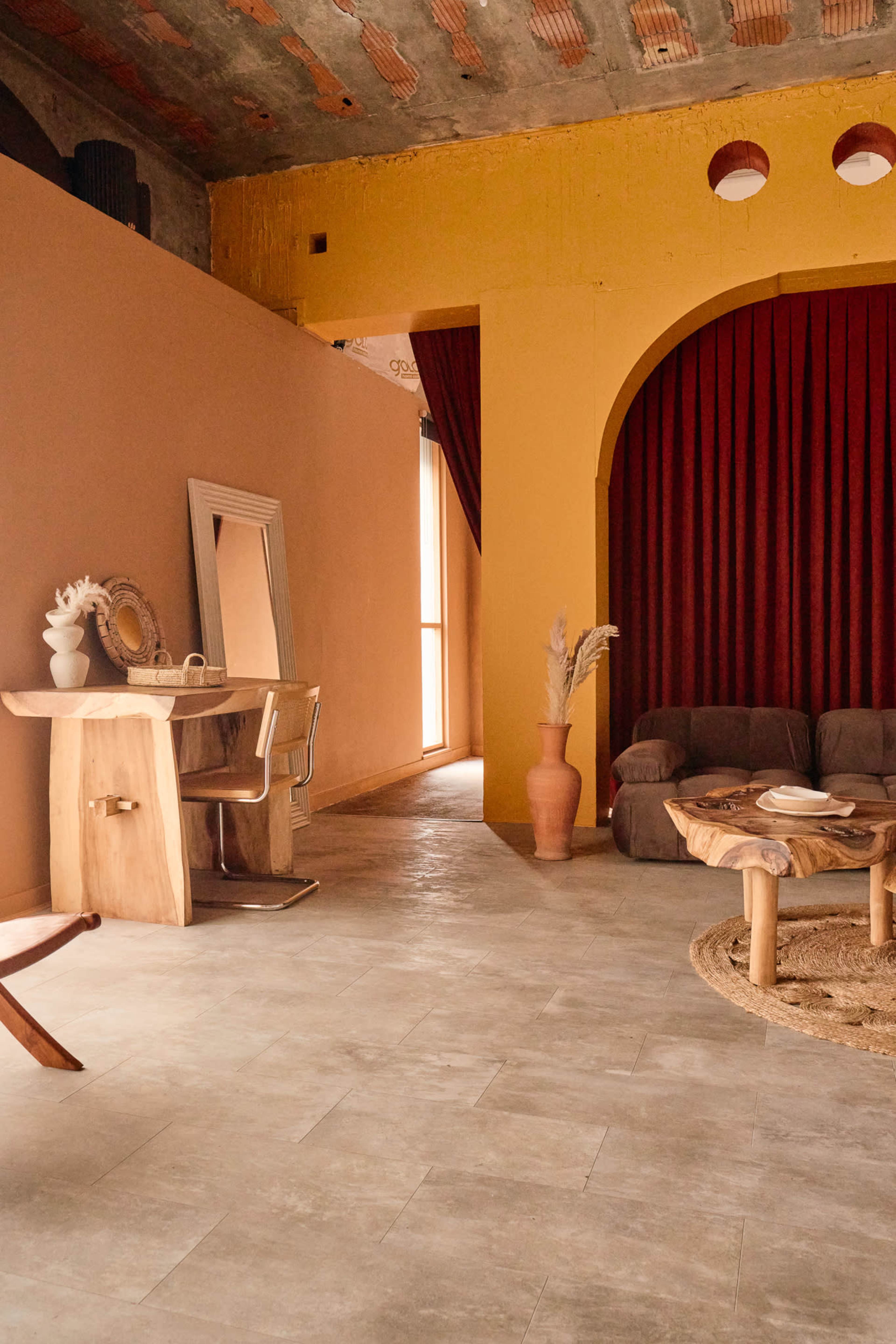 The image depicts a minimalist interior space featuring a wooden table, a mirror, a brown sofa, a round coffee table, and warm-toned walls with curtains.