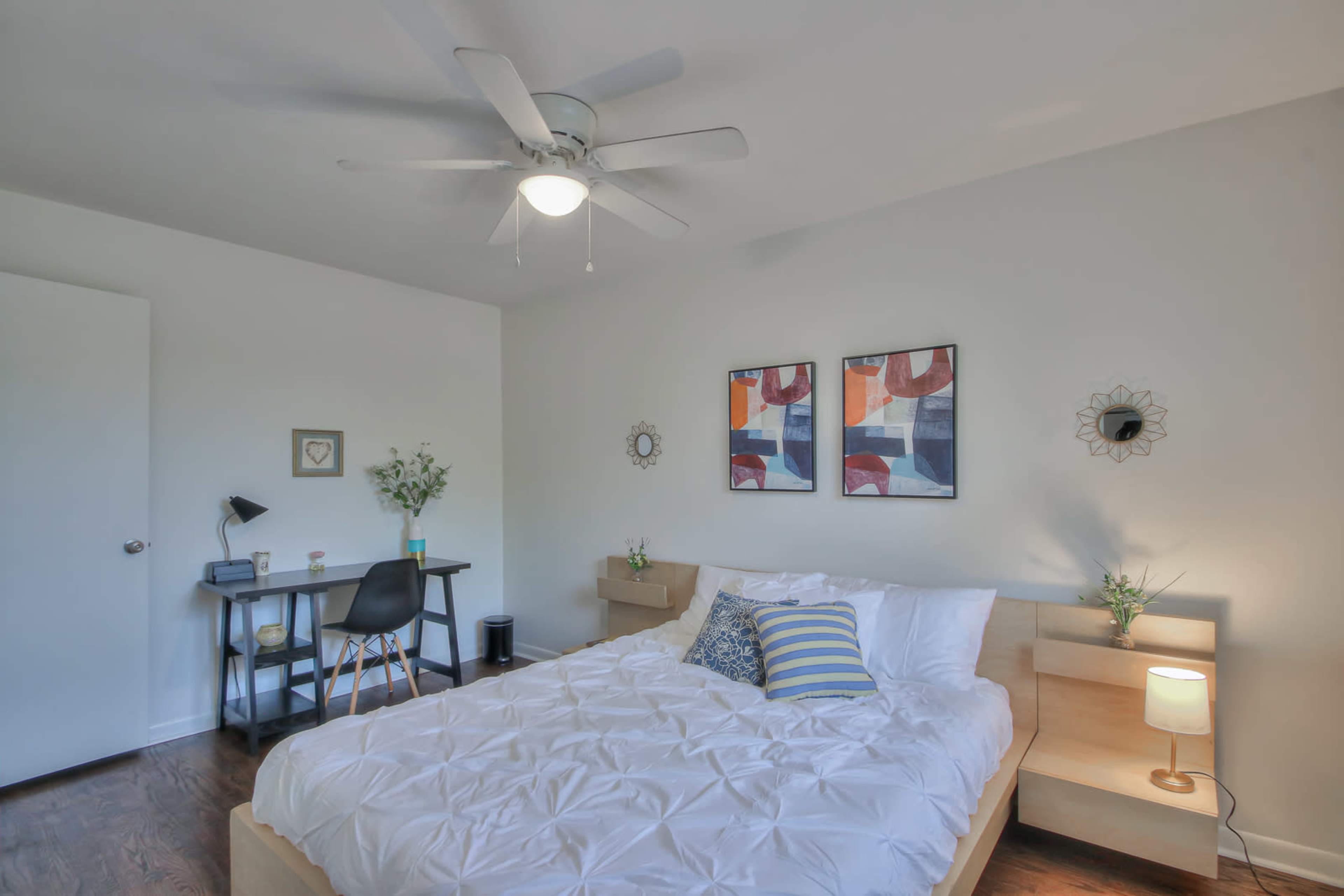 The image shows a modern bedroom featuring a bed, two decorative wall prints, a desk with a lamp, and a ceiling fan.