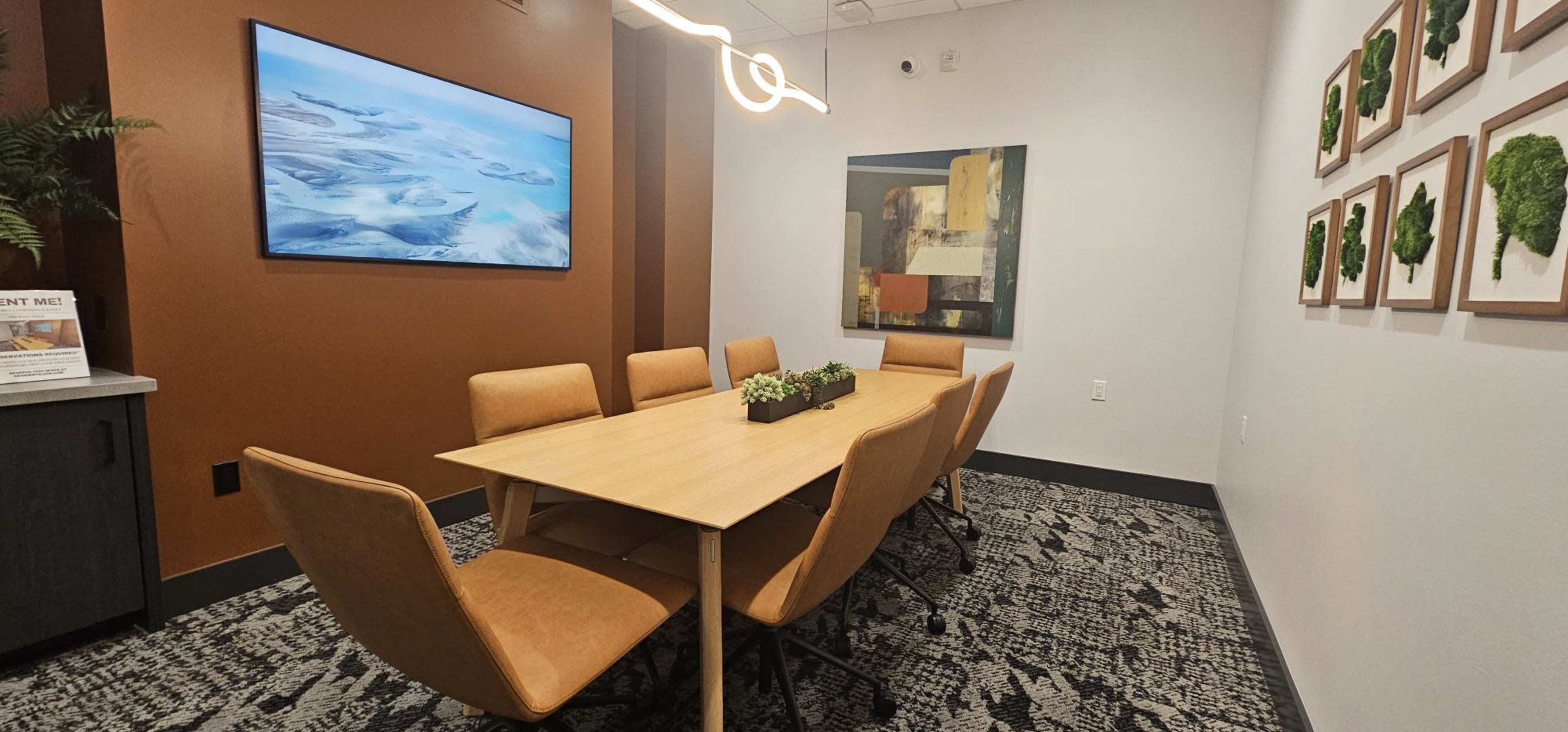 The image shows a modern conference room featuring a large table surrounded by six chairs, an abstract painting on the wall, and a television mounted on the opposite wall.