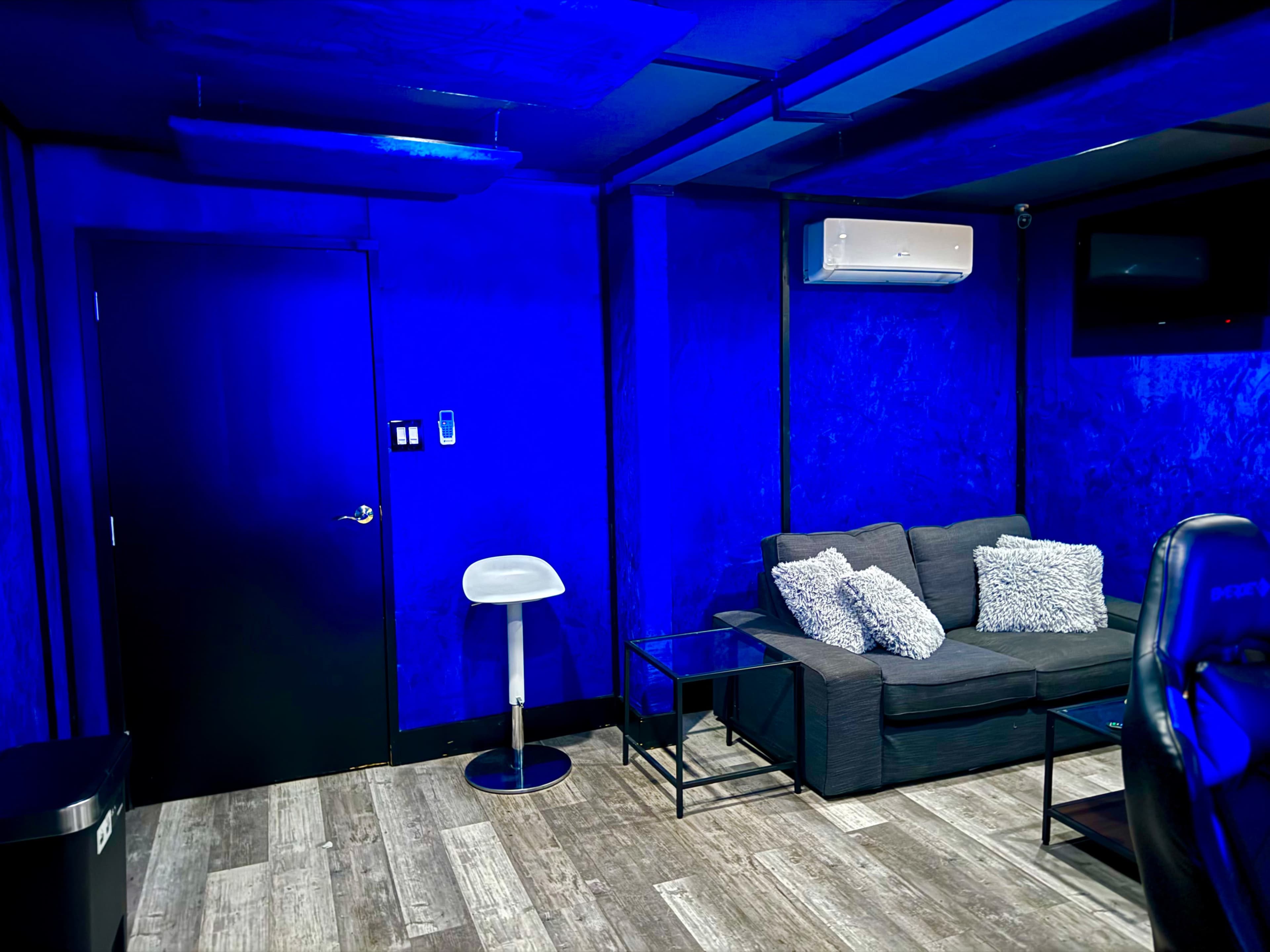 The room features blue walls, a black door, a gray sofa with pillows, a tall white stool, and a small black table.
