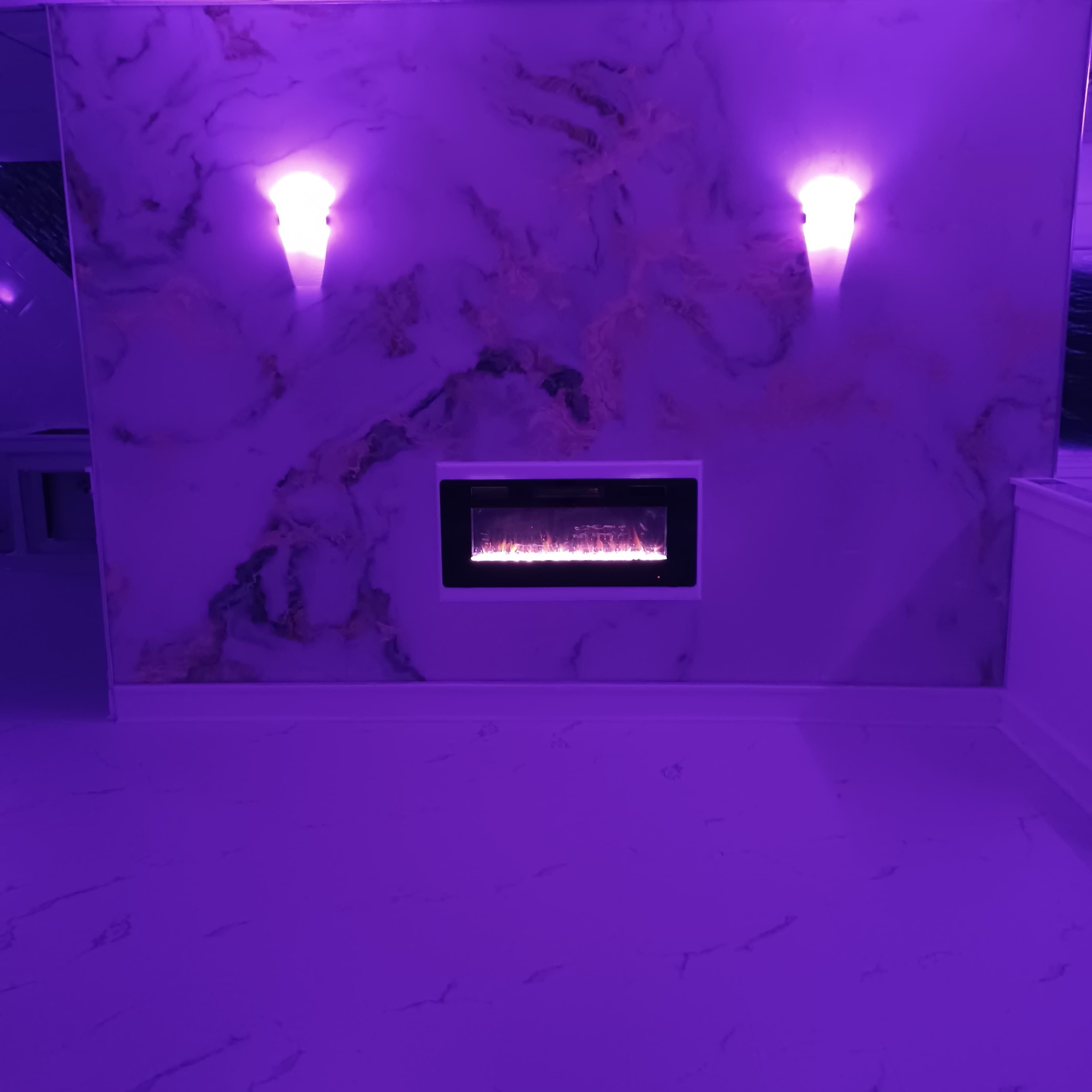 A room with a marble wall in shades of purple, featuring an electric fireplace centered below two wall sconces.