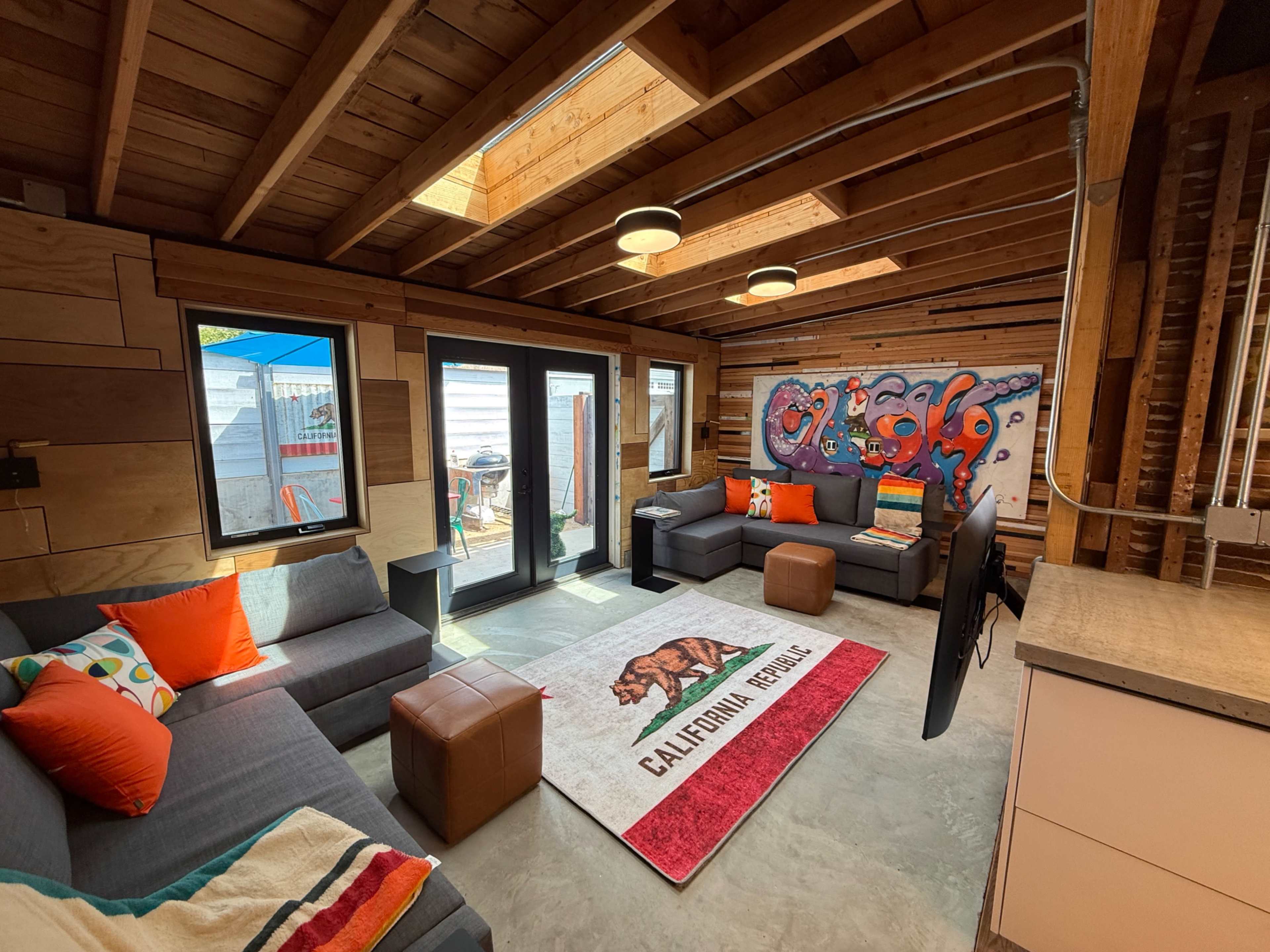 The image shows a modern living room with wooden beams, a colorful rug featuring a bear design, and a wall with vibrant graffiti art.