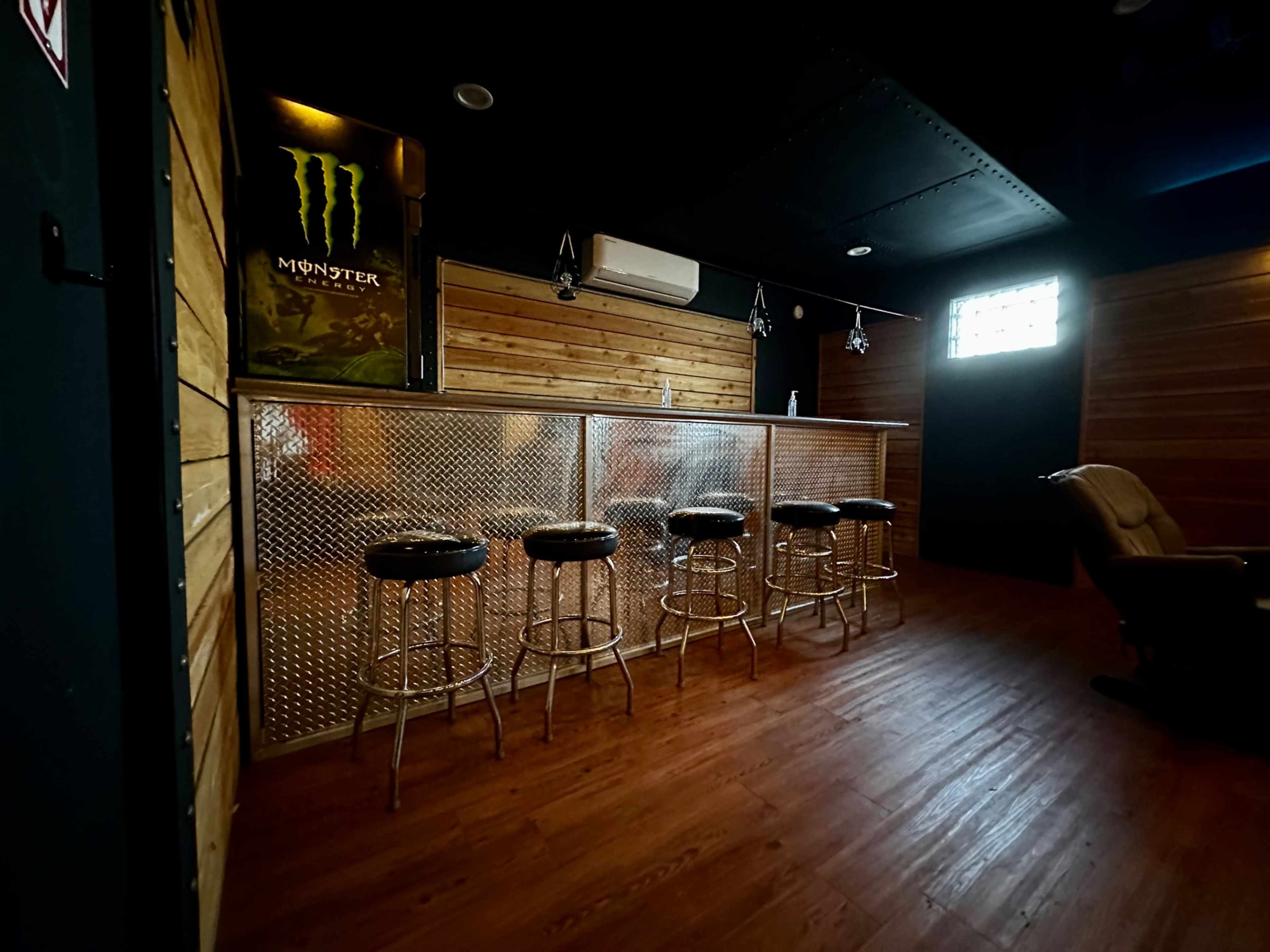 A dimly lit bar area with a metal counter and several high stools arranged in front of it, featuring wood-paneled walls.