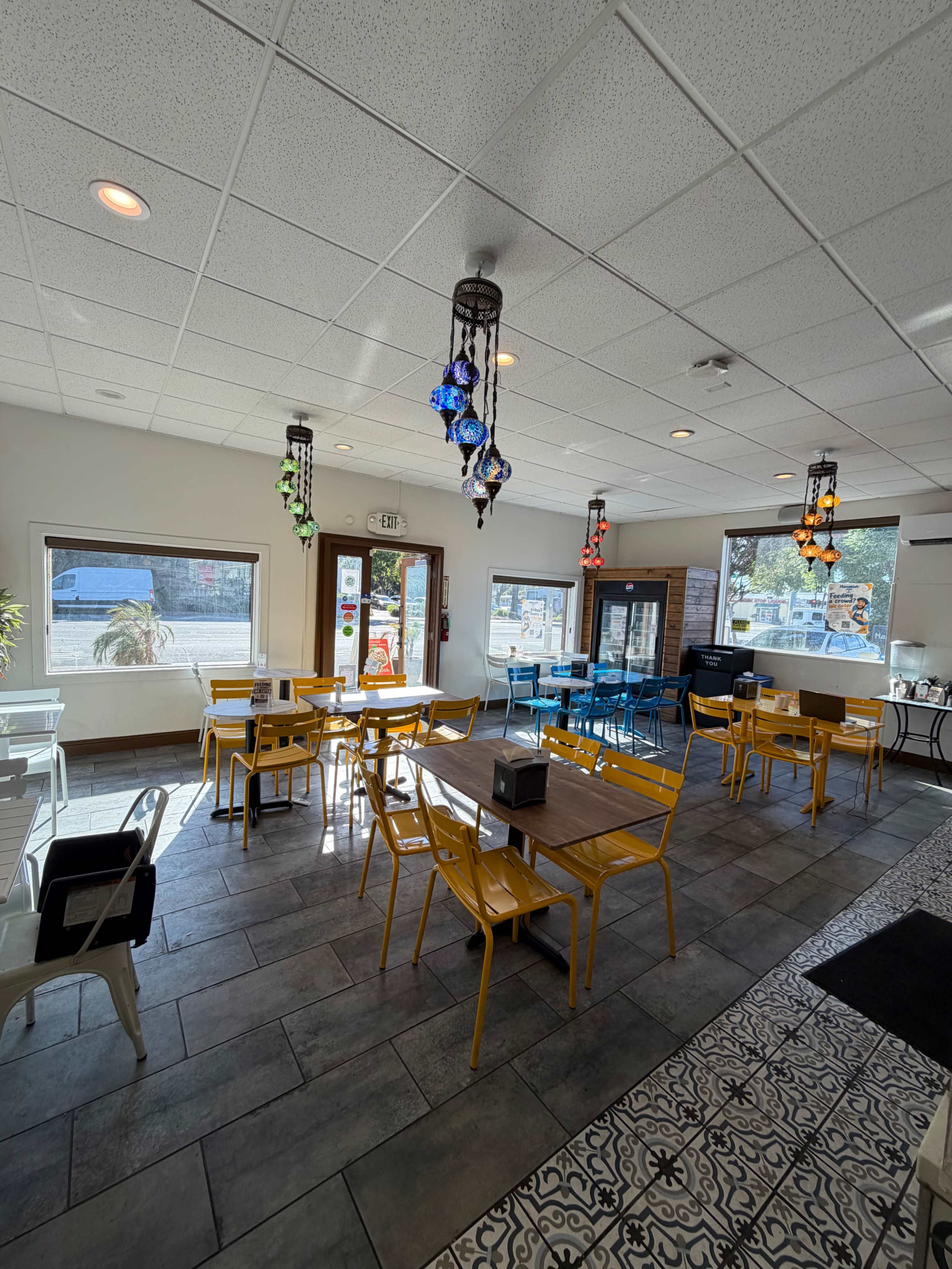 The image shows a brightly lit café interior with yellow and blue chairs arranged around several tables, and decorative hanging lanterns from the ceiling.