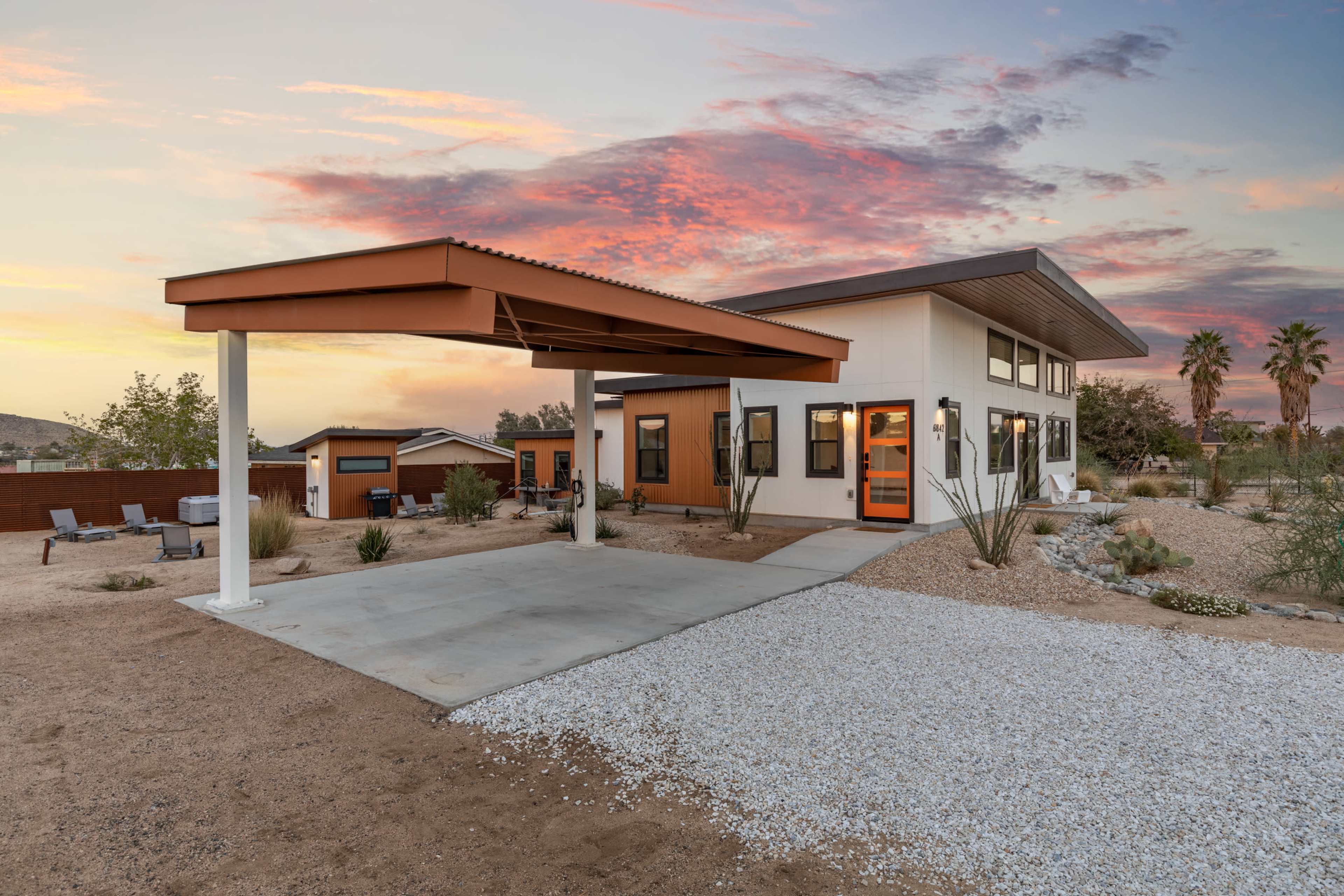 A modern house with a distinctive overhang and large windows is situated in a desert landscape at sunset.