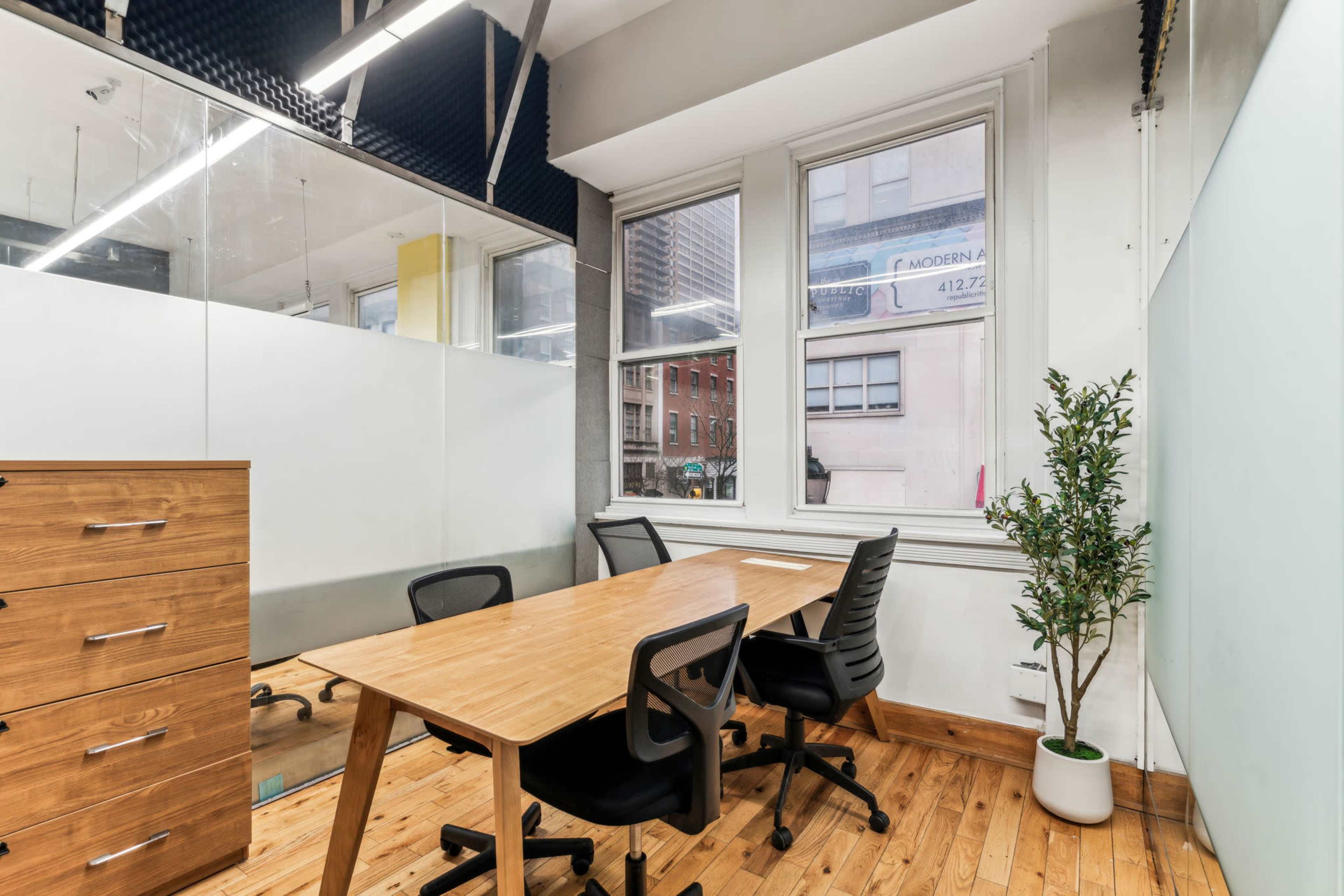 A modern office meeting room features a wooden table with four black chairs, a large window displaying urban buildings outside, and a small potted plant.