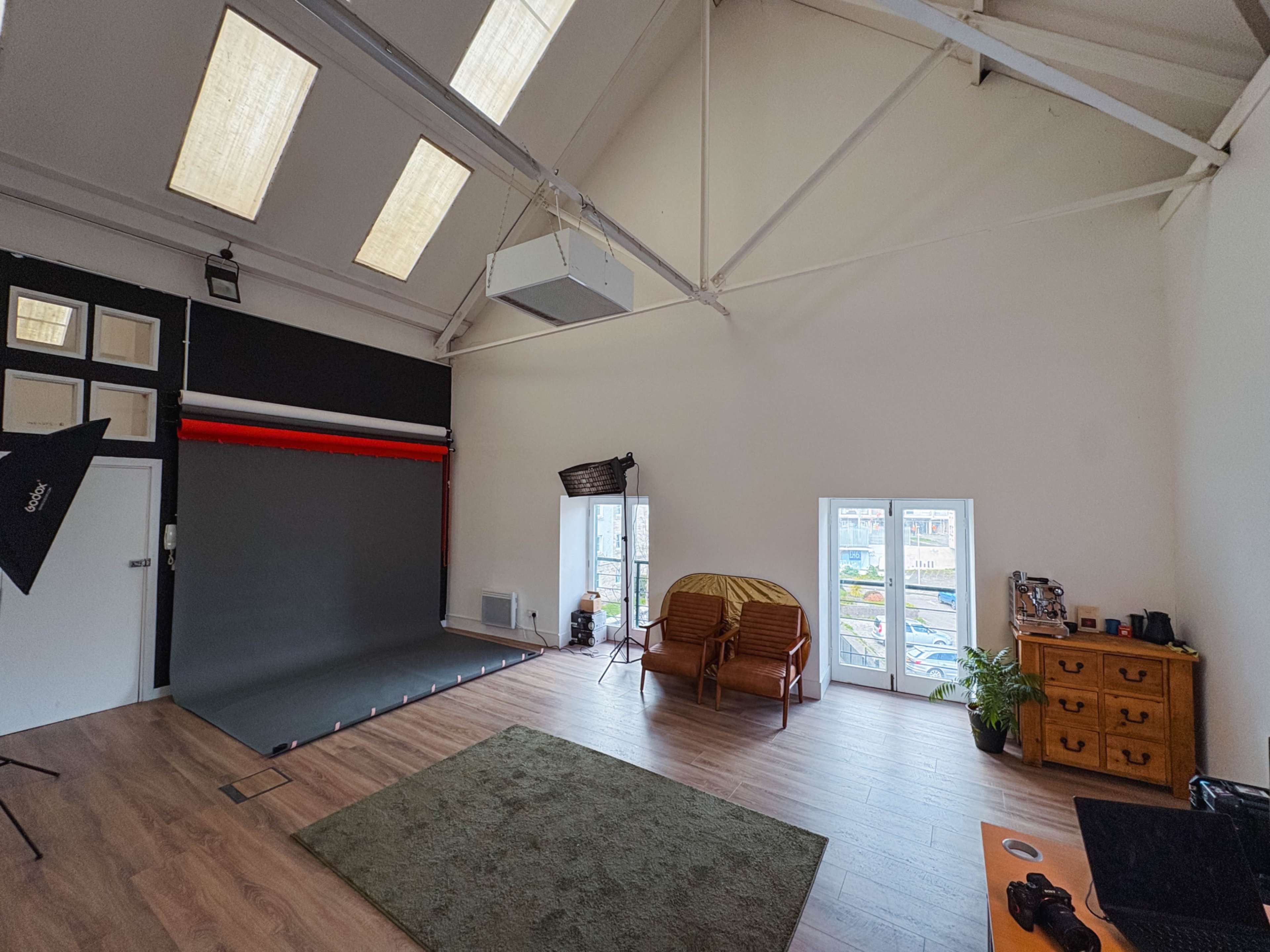 The image shows a spacious photo studio with a green area rug, a backdrop setup, and minimalist furniture including a chair and a potted plant.