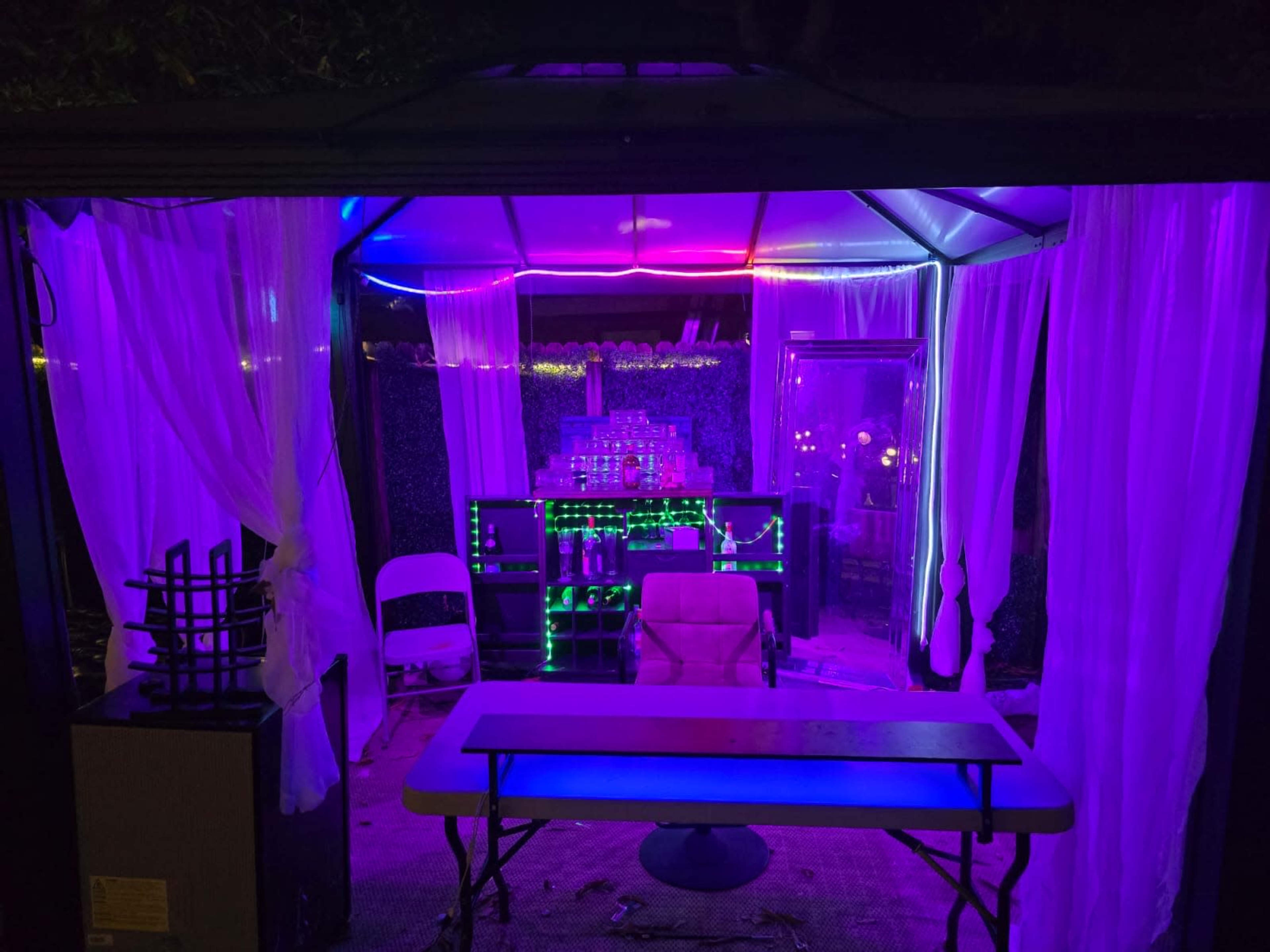 The image shows a dimly lit outdoor gazebo decorated with colorful LED lights and sheer curtains, featuring a table and chairs inside.