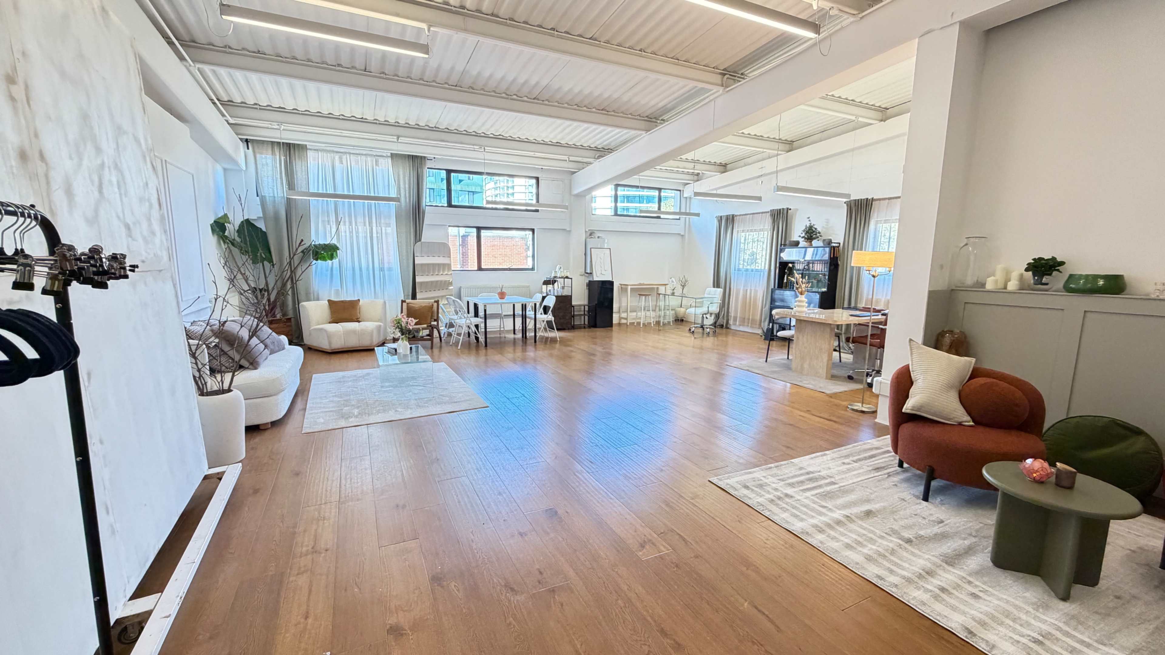 The image shows a spacious, well-lit interior with wooden flooring, featuring various seating areas, a dining setup, and decorative plants throughout.