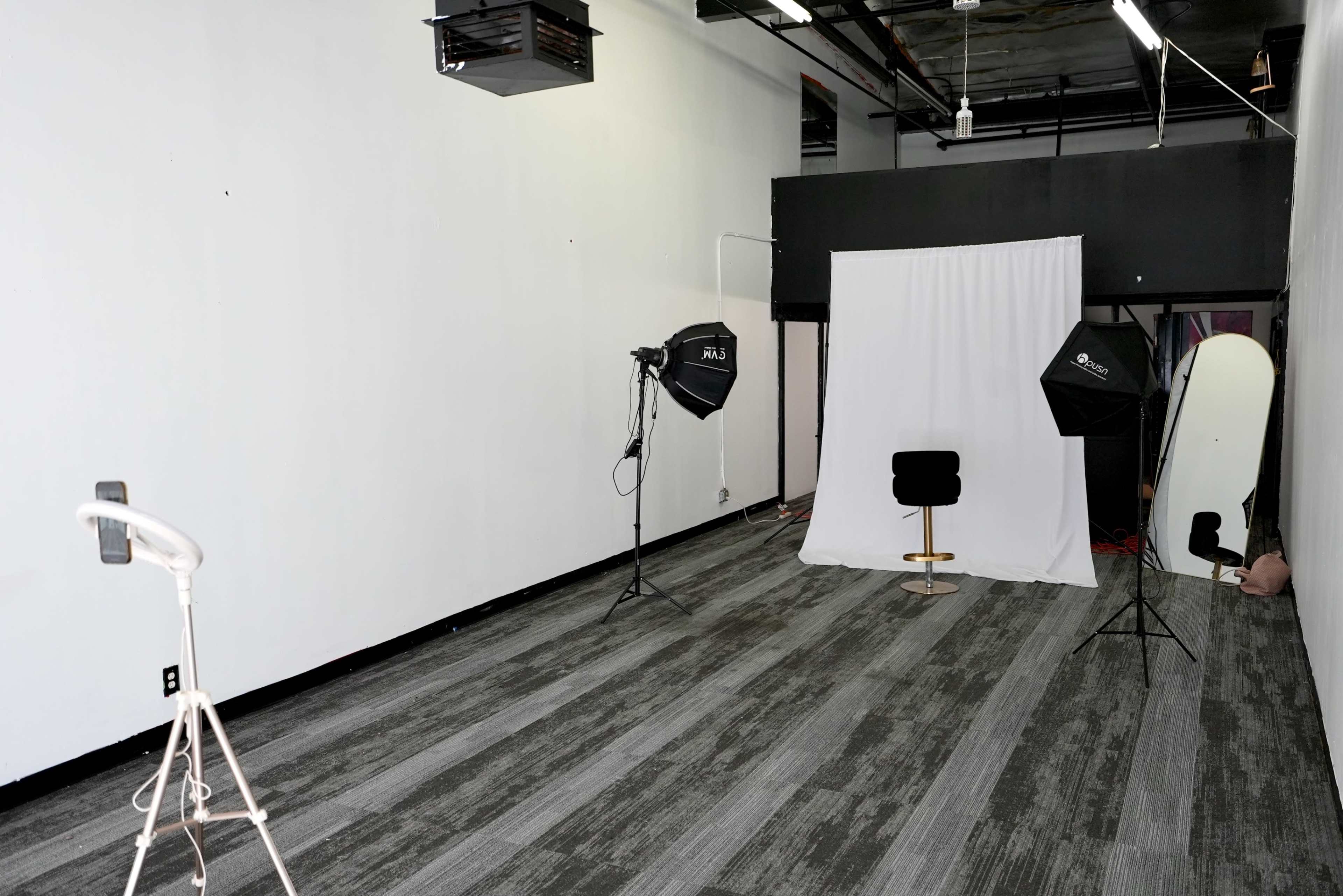 The image shows a vacant studio space with a white backdrop, two lighting setups, and a black chair positioned on a gray carpet.
