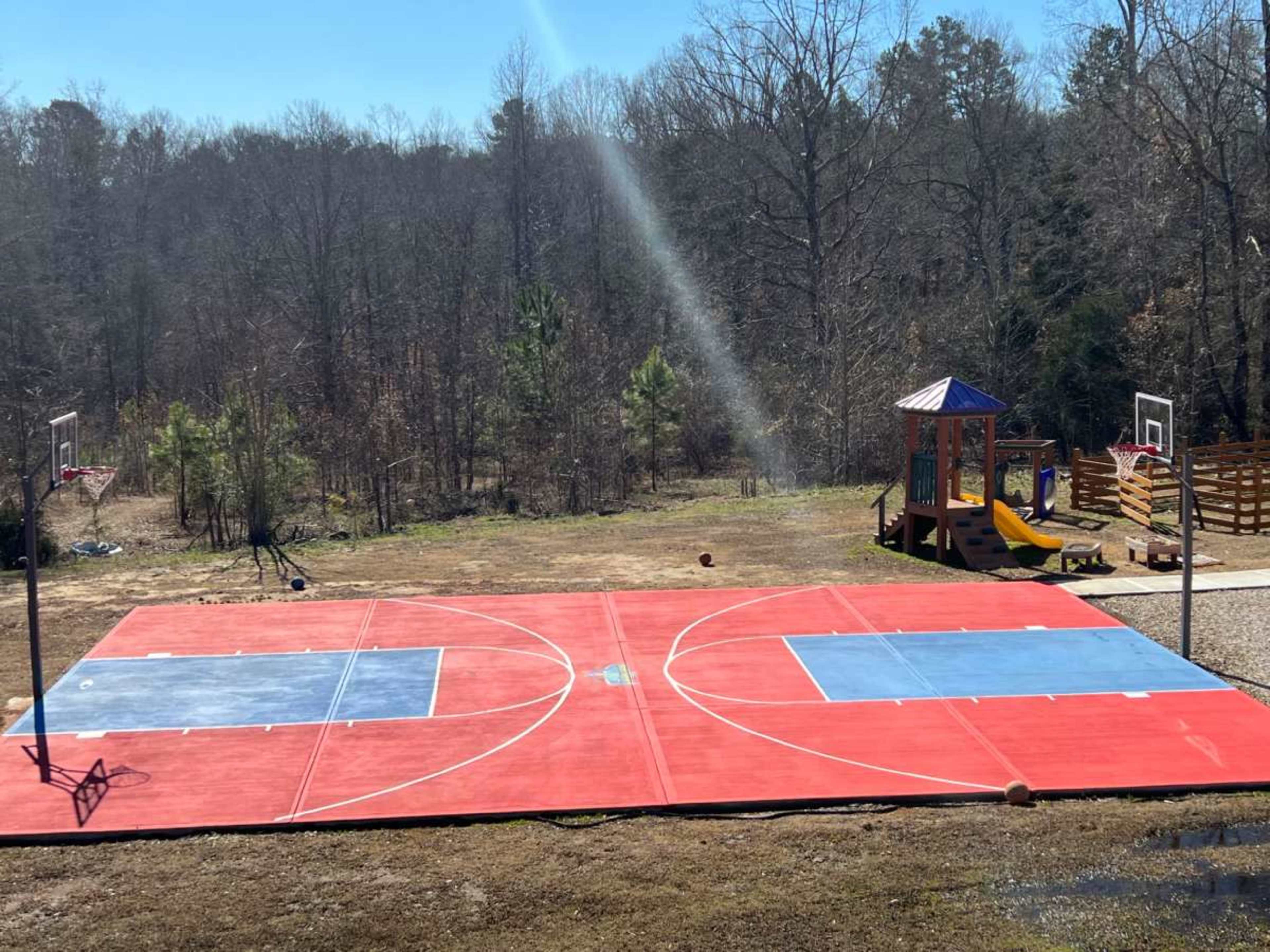 The image shows a newly painted outdoor basketball court with two hoops and a playground structure nearby.