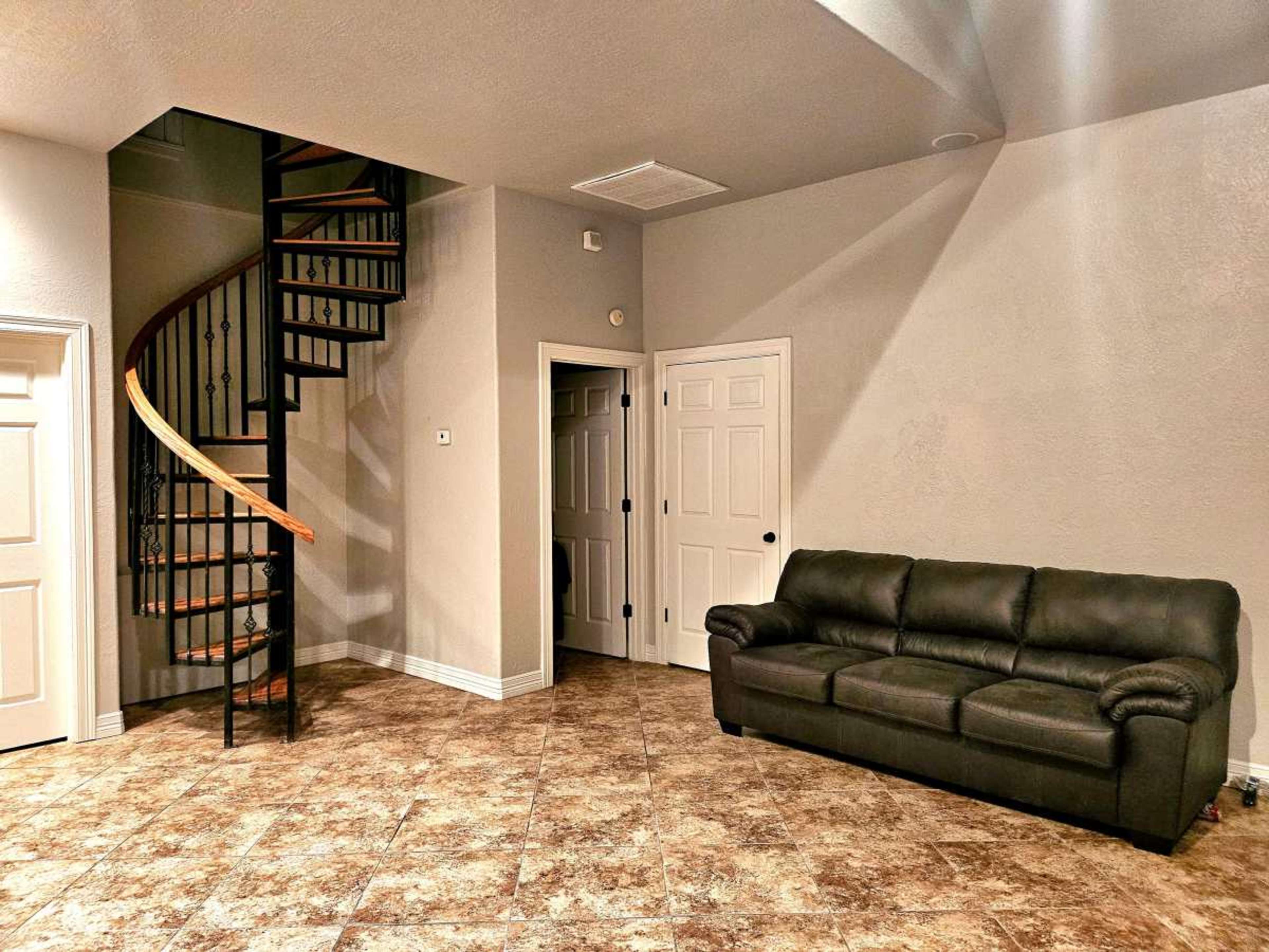 The image features a room with a spiral staircase in one corner, a dark couch against the wall, and a light-colored wall with several doors.
