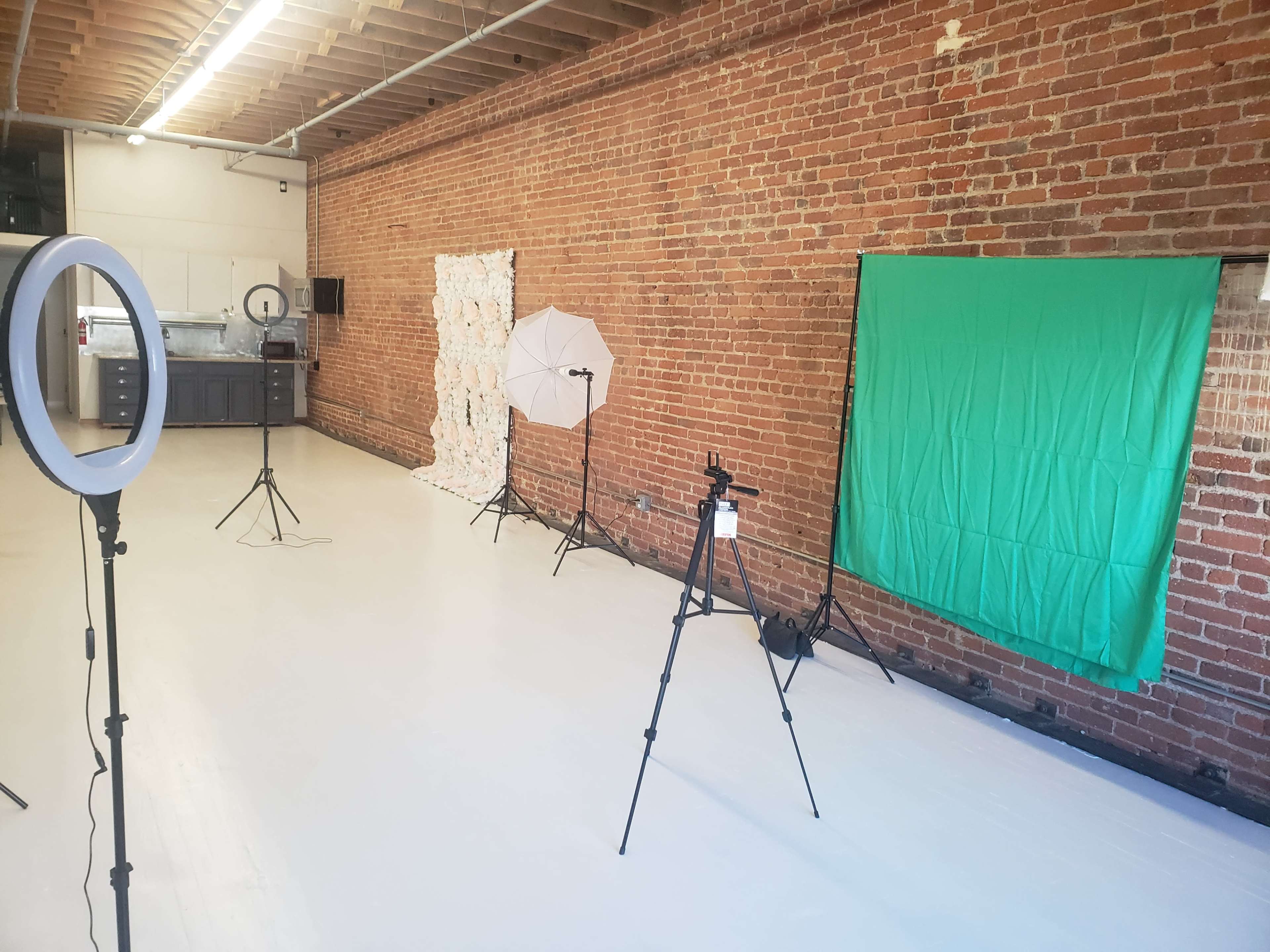 The image shows a spacious room with exposed brick walls and white flooring, featuring a green screen, lighting equipment, and a backdrop setup for photography or videography.