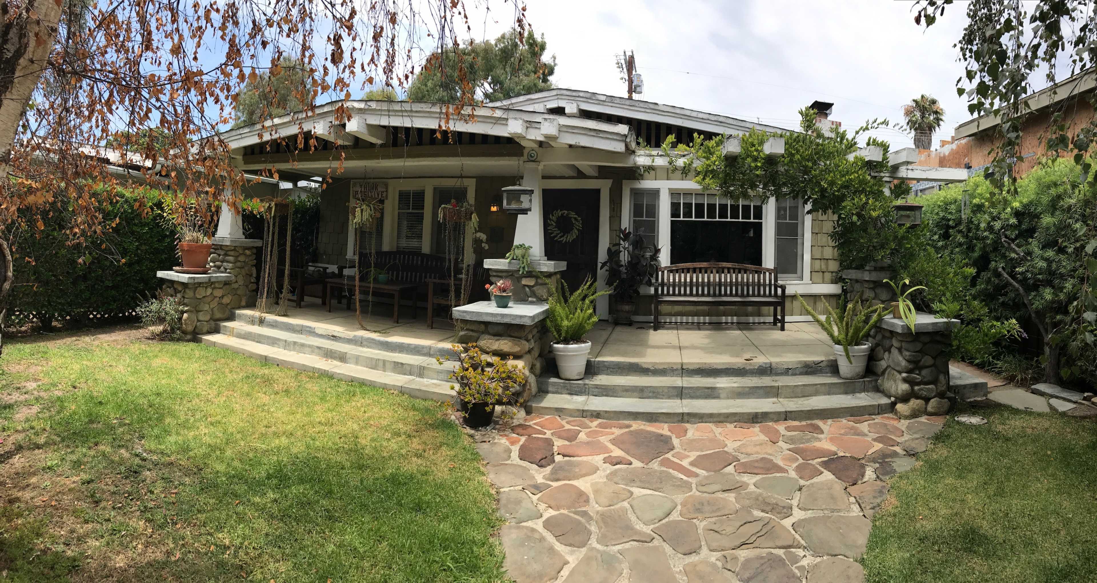 The image shows a single-story house with a wide porch, surrounded by a landscaped yard featuring stone pathways and various plants.