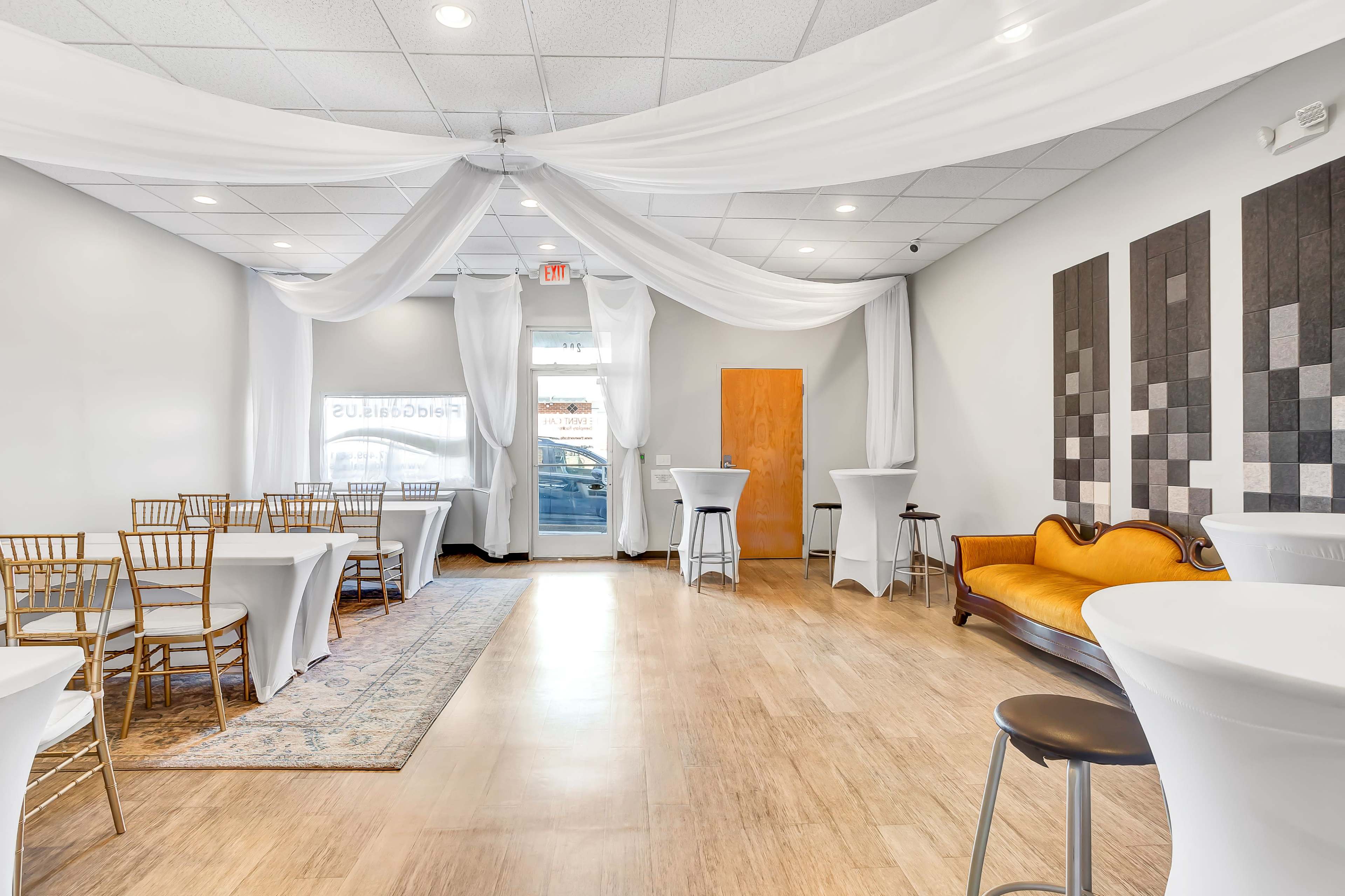 An interior space with white drapery, a few high-top tables, gold chiavari chairs, a vintage sofa, and a clean wooden floor.