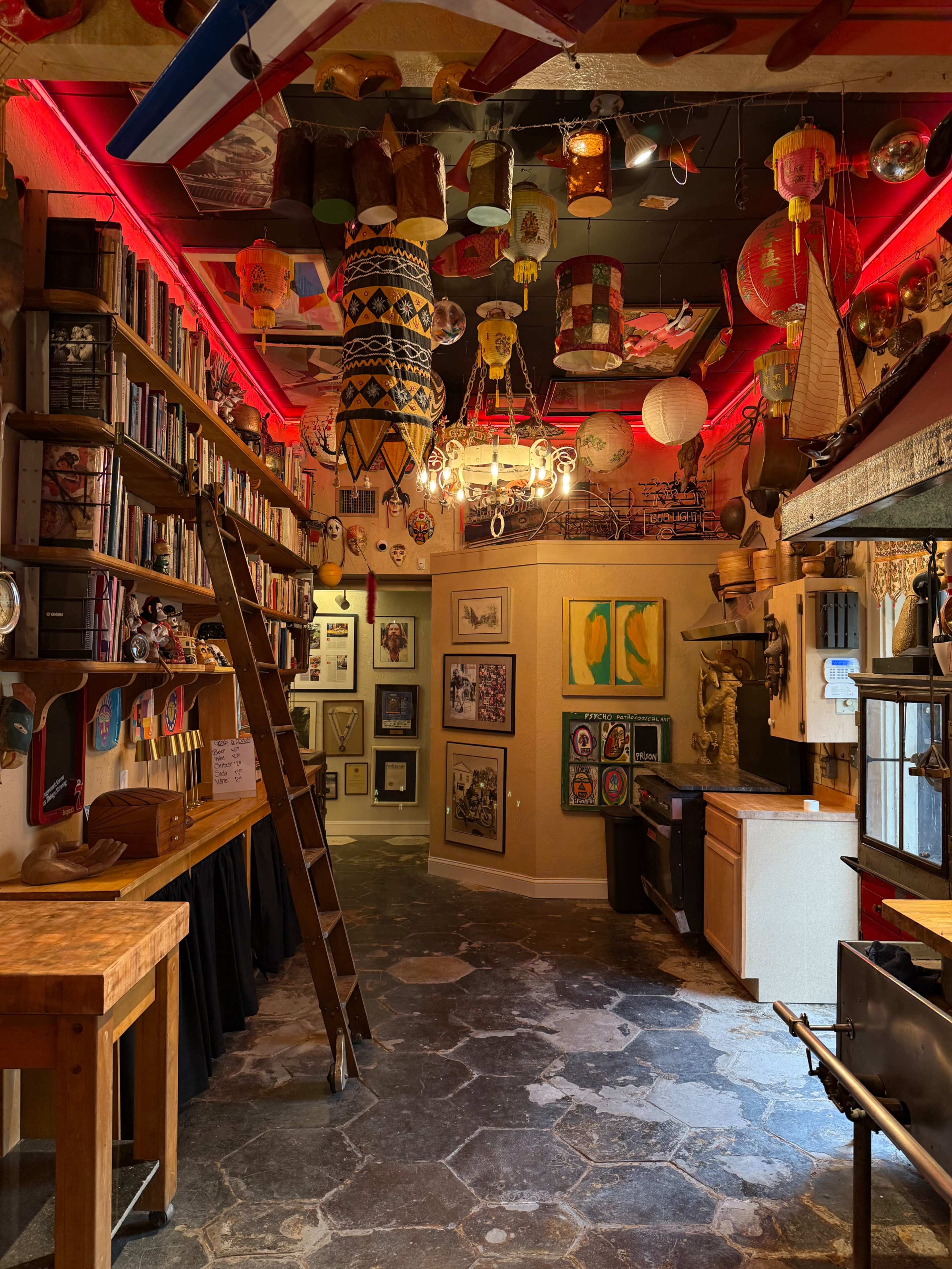 The image shows a vibrant, eclectic interior space filled with various decorative objects, artworks, and bookshelves, along with a wooden ladder leaning against the wall.