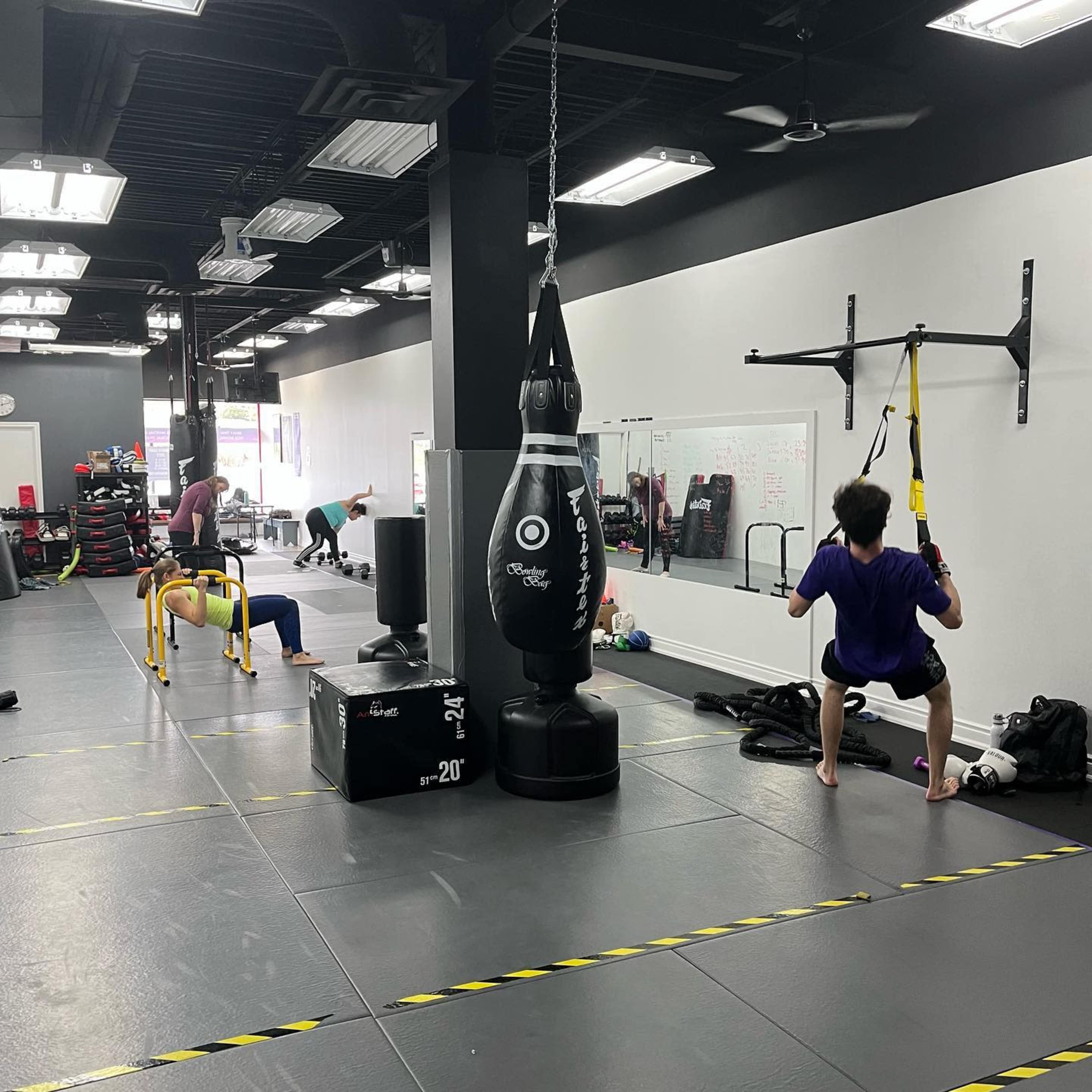 A gym interior features several workout stations, including a punching bag, TRX suspension trainer, and individuals exercising with various equipment.