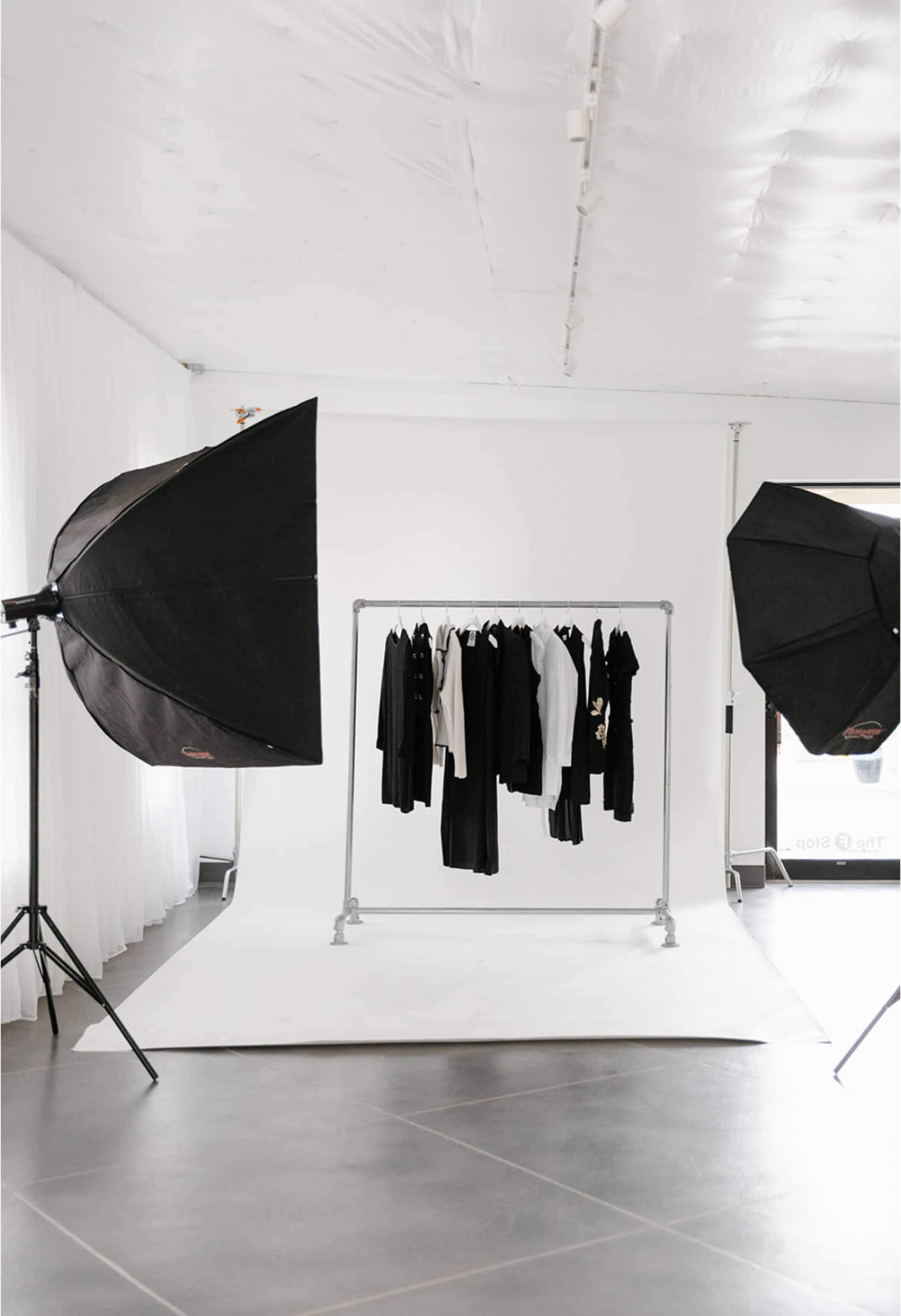 A clothing rack with various garments hangs in a bright studio space, flanked by two large softbox lights.