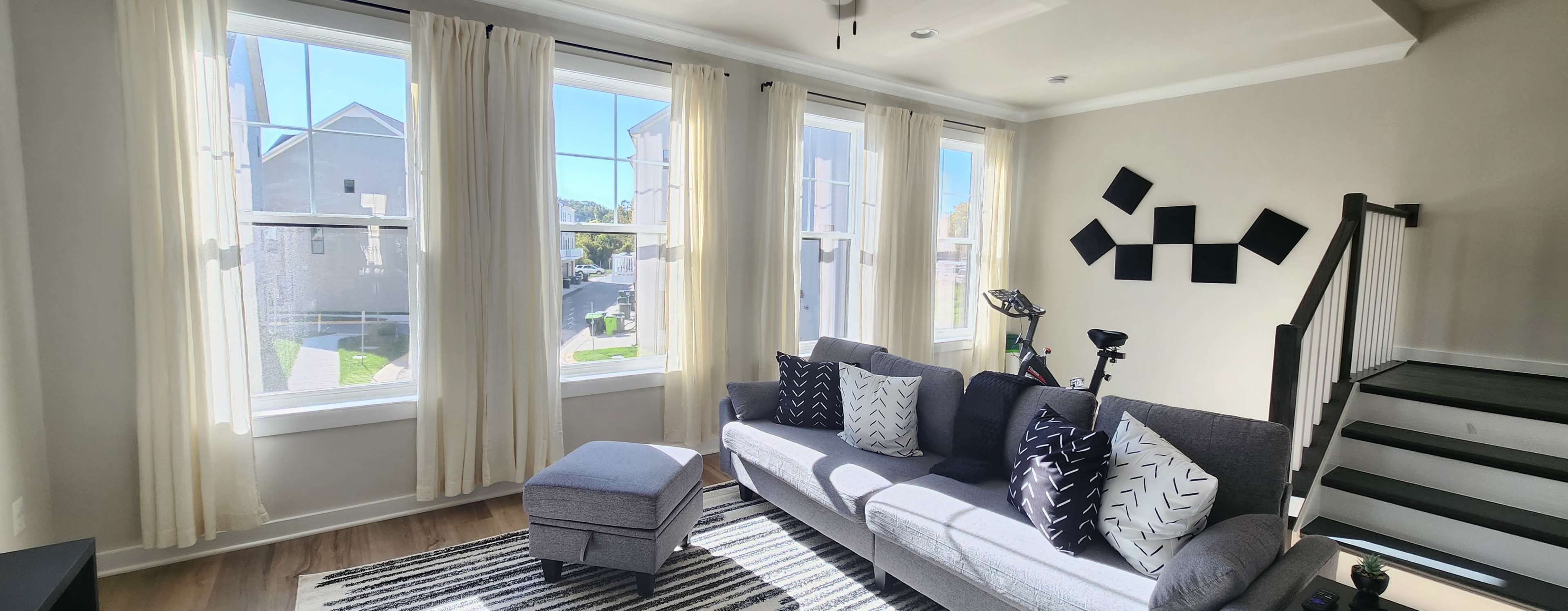 A bright living room with large windows, a gray sofa adorned with black and white pillows, an ottoman, a striped rug, and a staircase in the background.