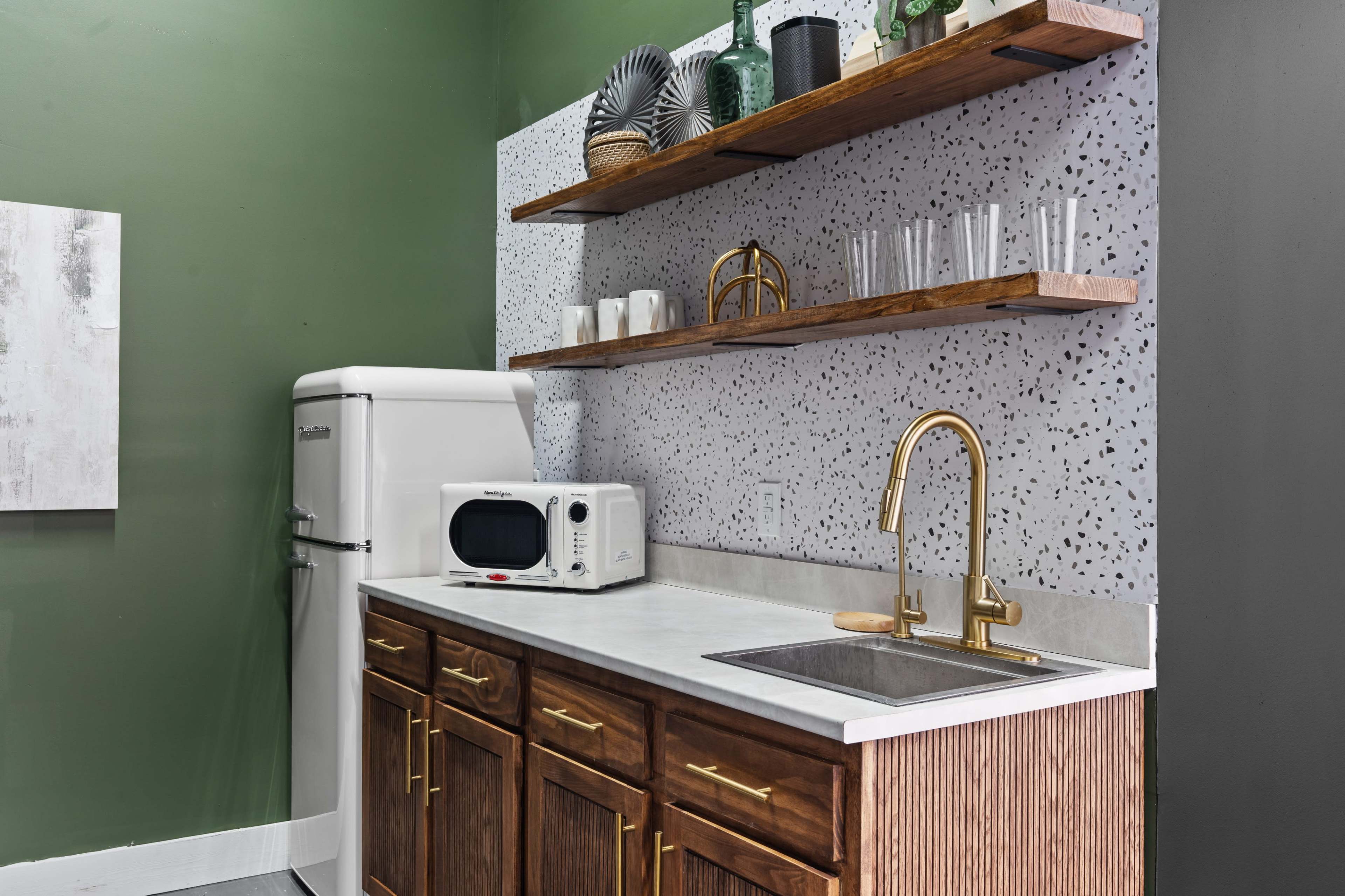 The image shows a modern kitchenette with a wooden countertop, a white refrigerator, a microwave, and open shelves displaying various kitchenware against a green wall.