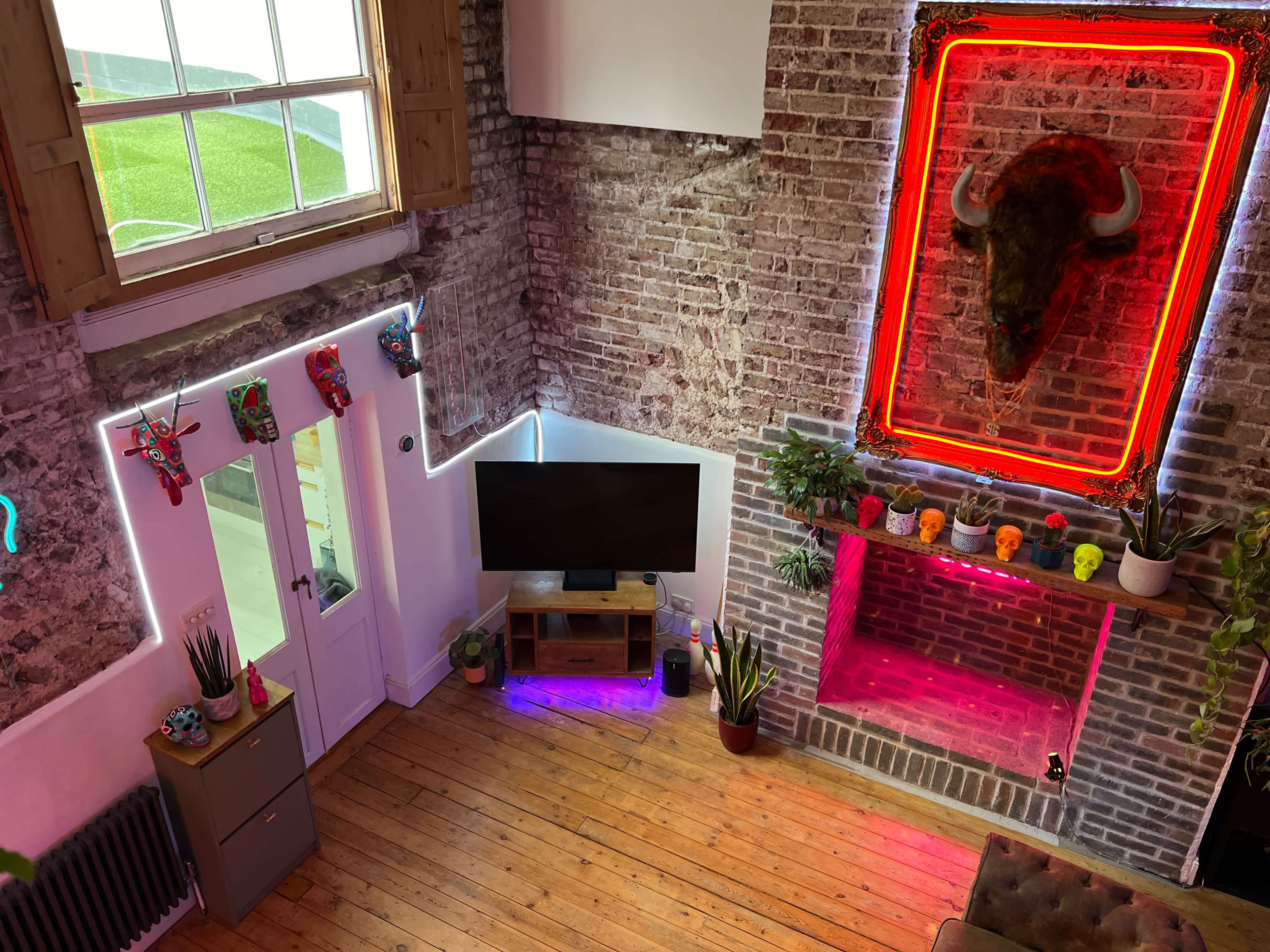 A modern living space with exposed brick walls, a television on a wooden stand, colorful decorations, and a neon-lit bull's head mounted above a fireplace.
