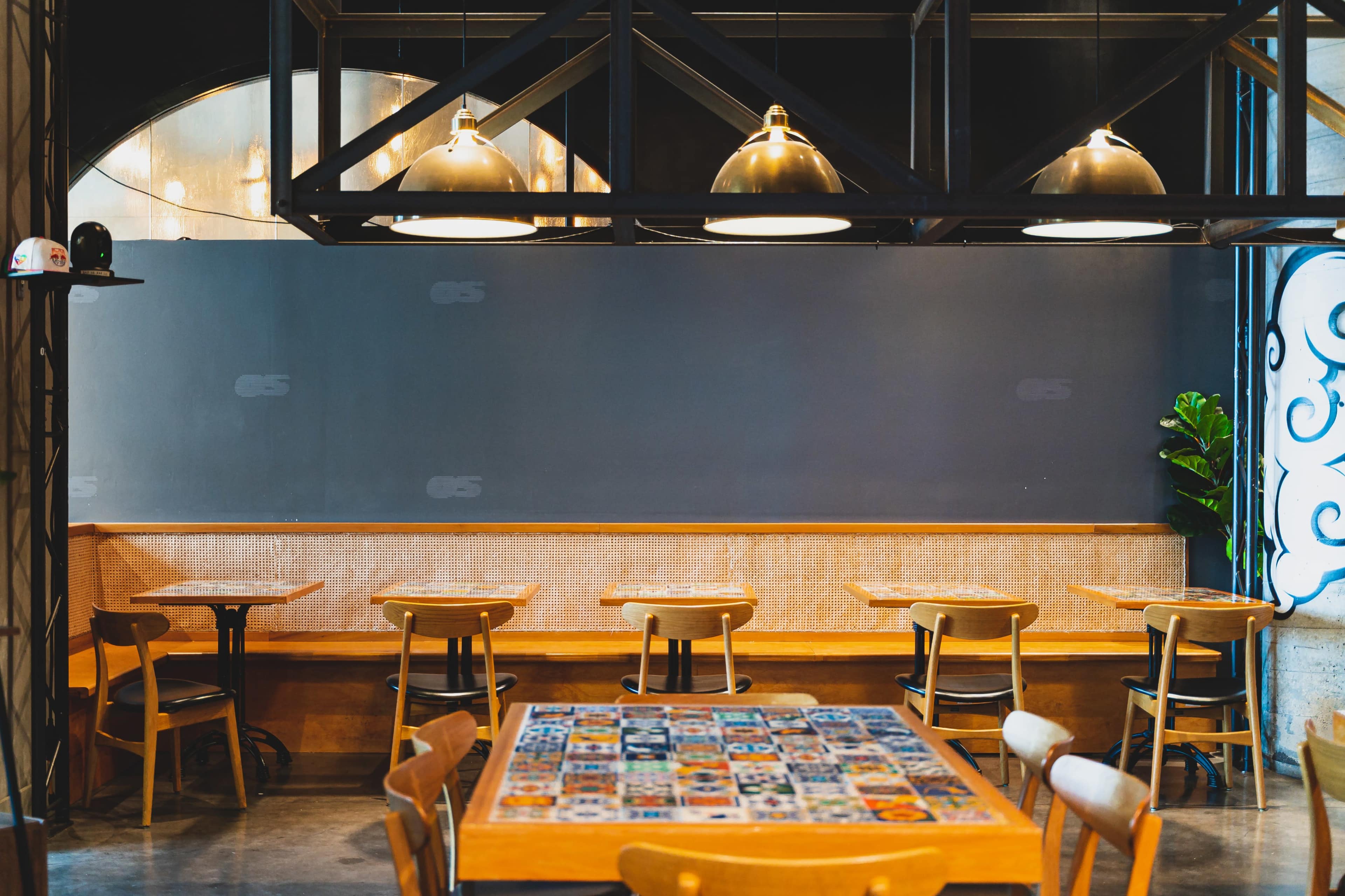 A modern cafe interior with wooden tables and chairs arranged around a central table decorated with various coasters, and a dark wall in the background.