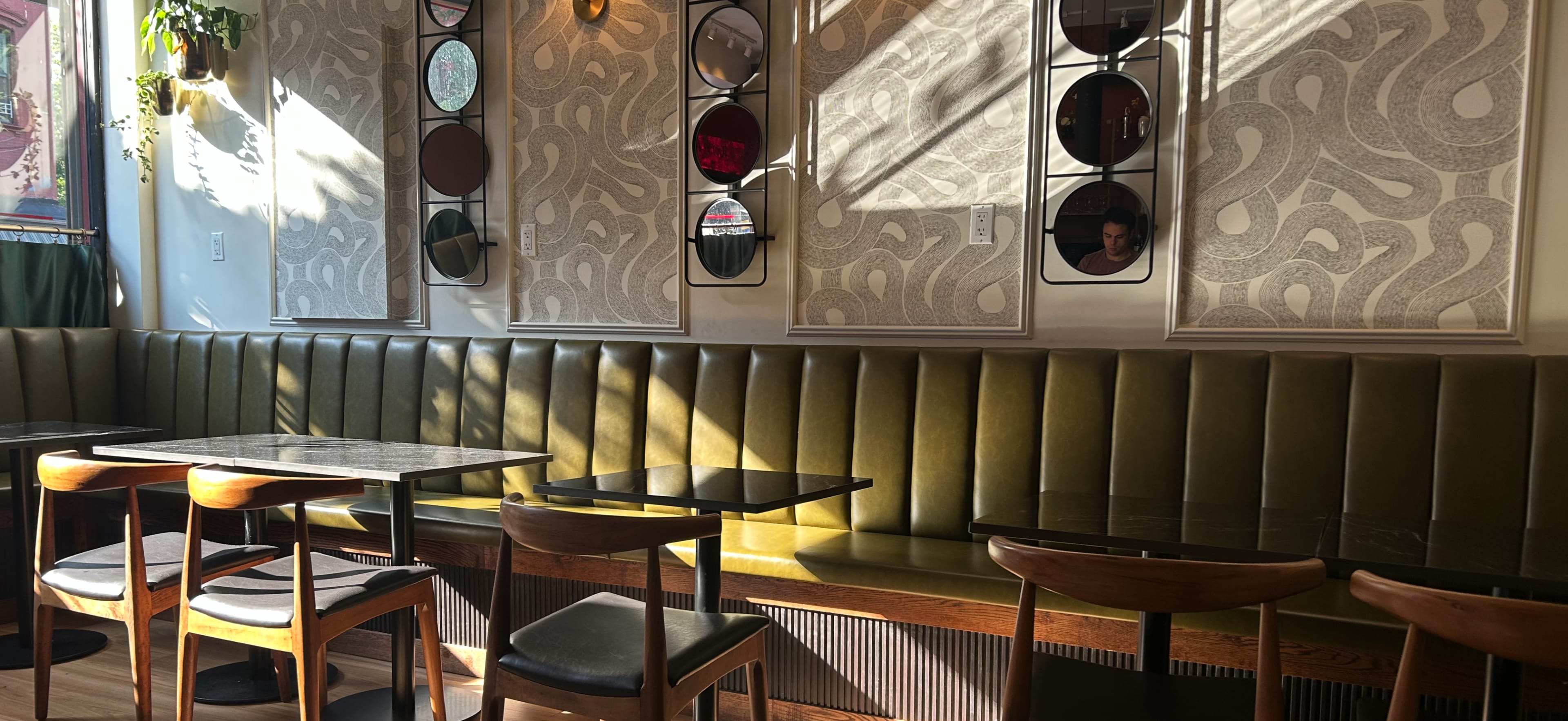 The image shows an interior of a café with green upholstered benches, wooden tables, and patterned wall decor, illuminated by sunlight streaming through the windows.