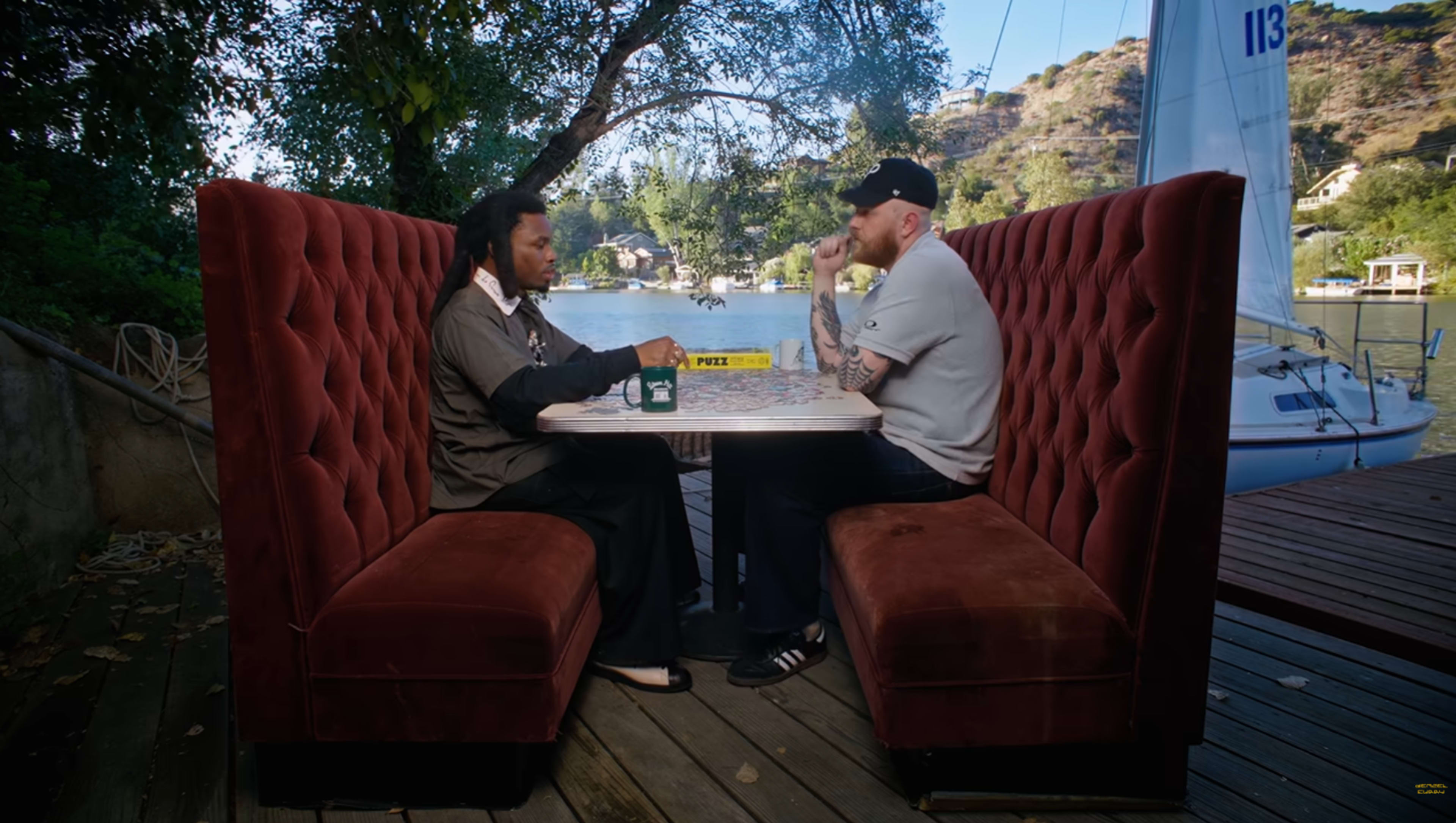 Two people sit at a small table in a secluded outdoor booth overlooking a calm waterway, with a sailboat in the background.