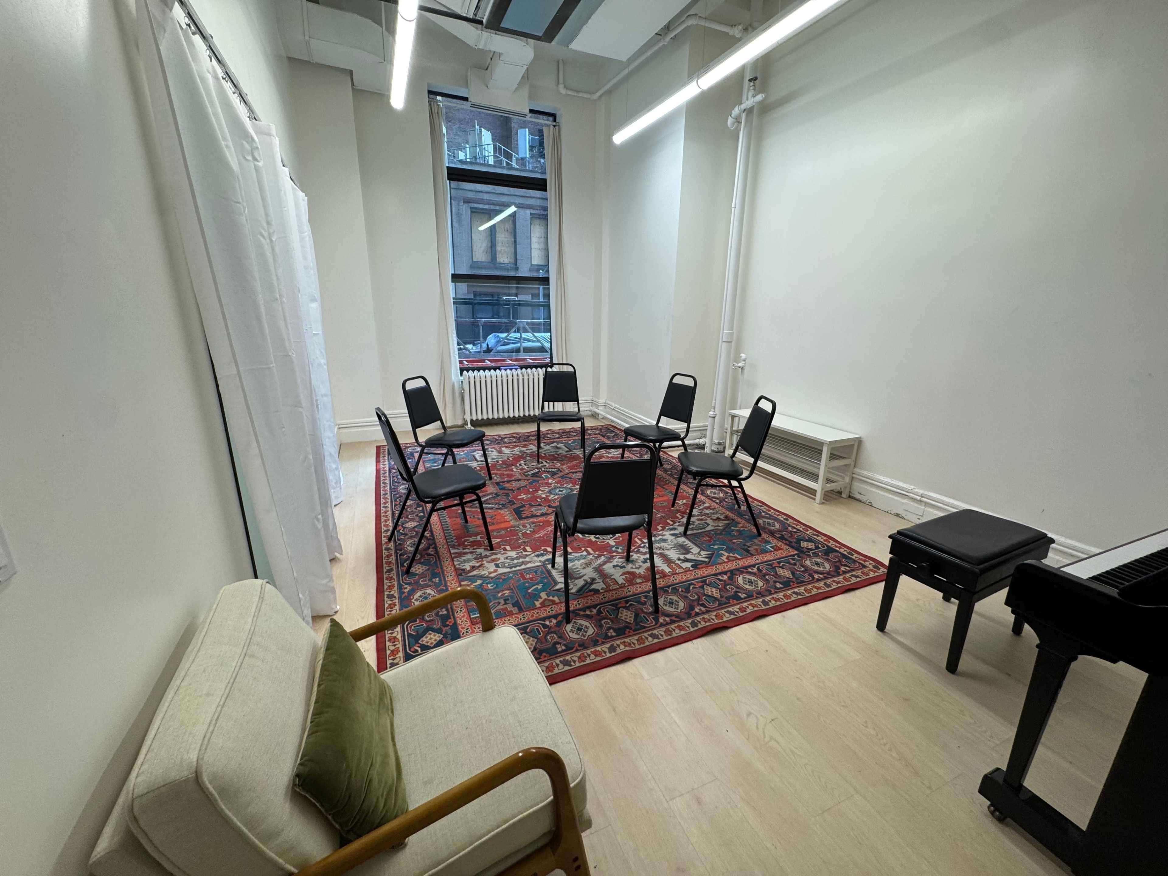 The image shows a small, equipped room with a patterned rug, several chairs arranged in a circle, a couch, and a piano in one corner.