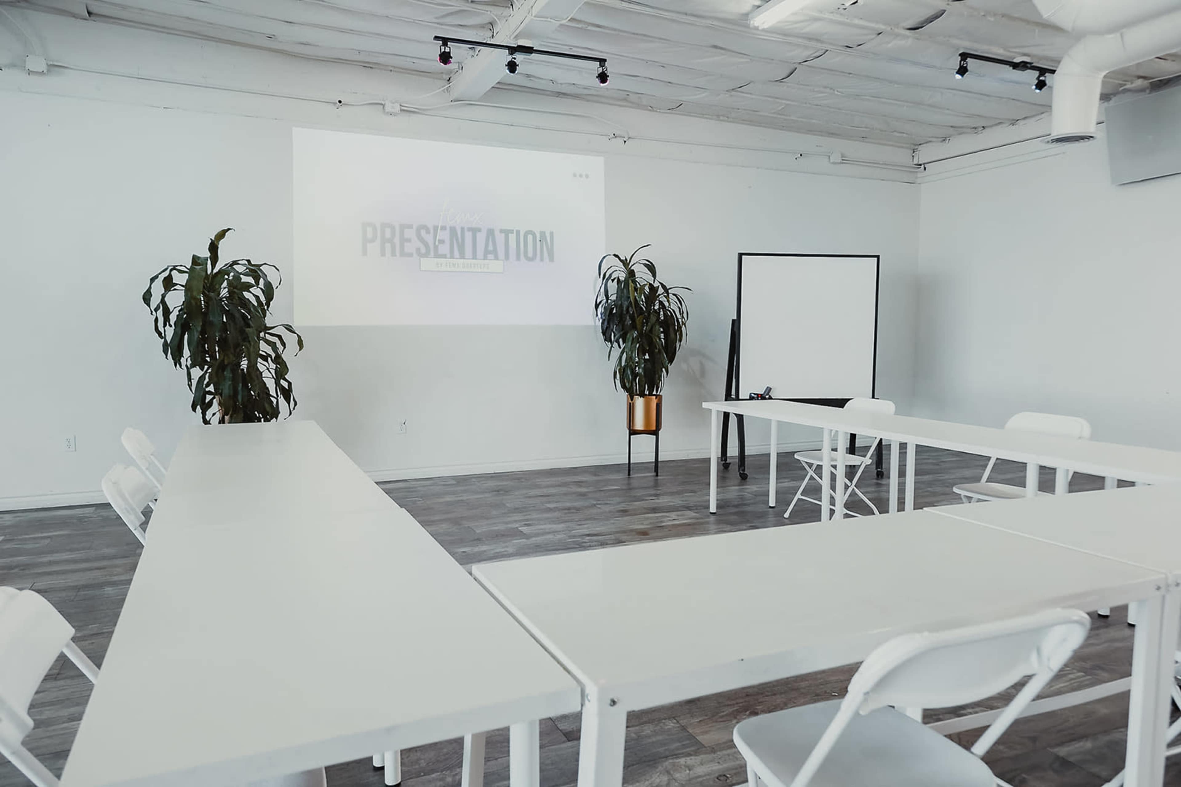 A bright meeting room features white tables arranged in a U-shape, two potted plants, and a projector screen displaying the word "PRESENTATION."