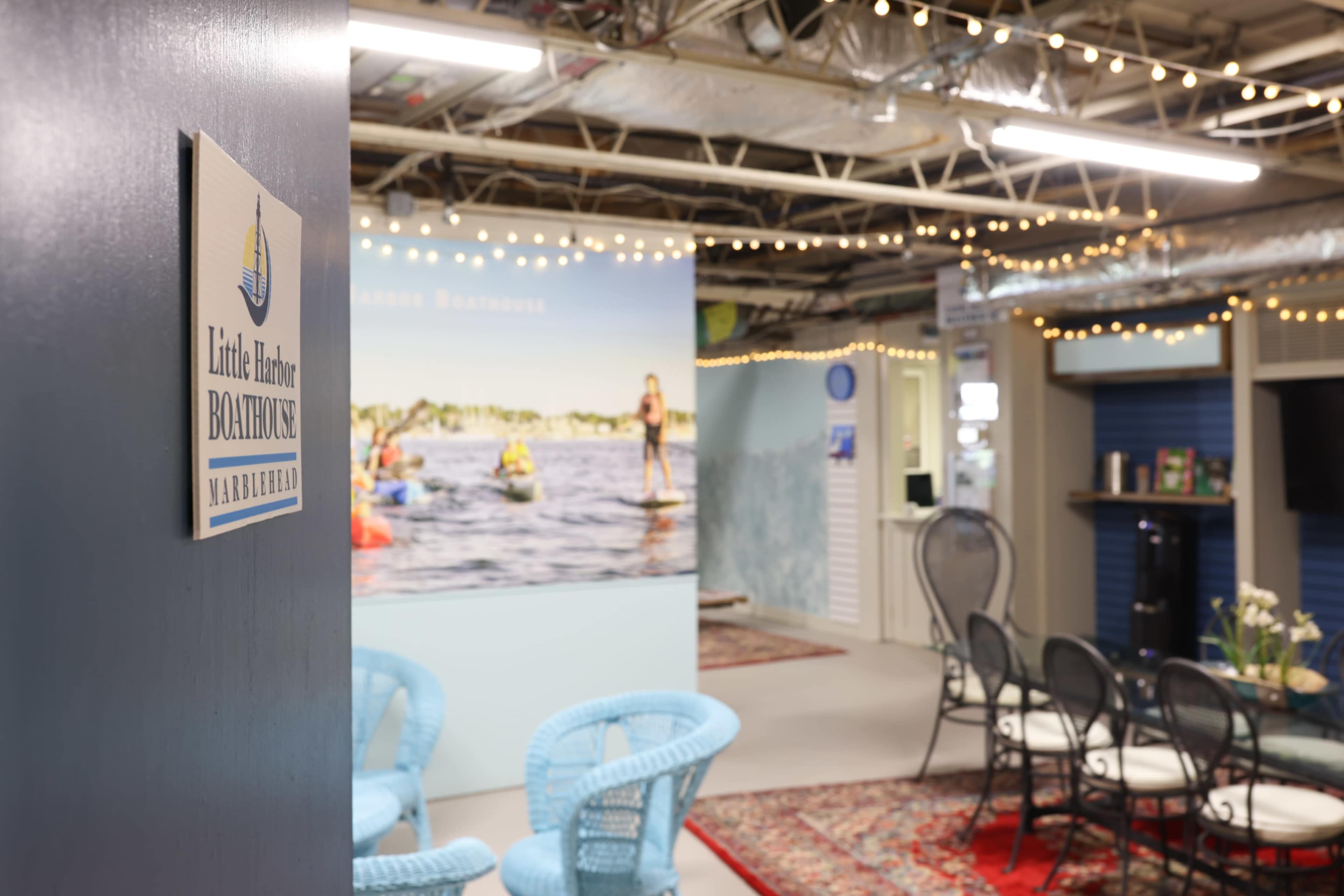 The interior of a boathouse facility with a sign for "Little Harbor Boathouse," featuring seating areas, decorative lighting, and a backdrop of people kayaking.