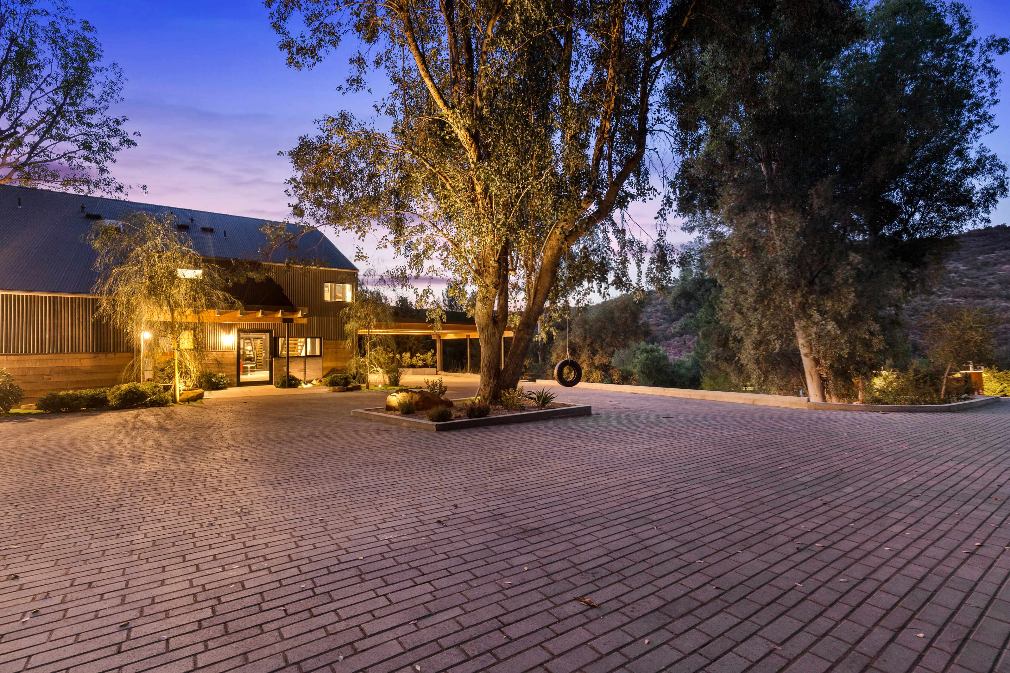 A spacious brick-paved area at dusk features a large structure with a wooden facade and surrounding trees.