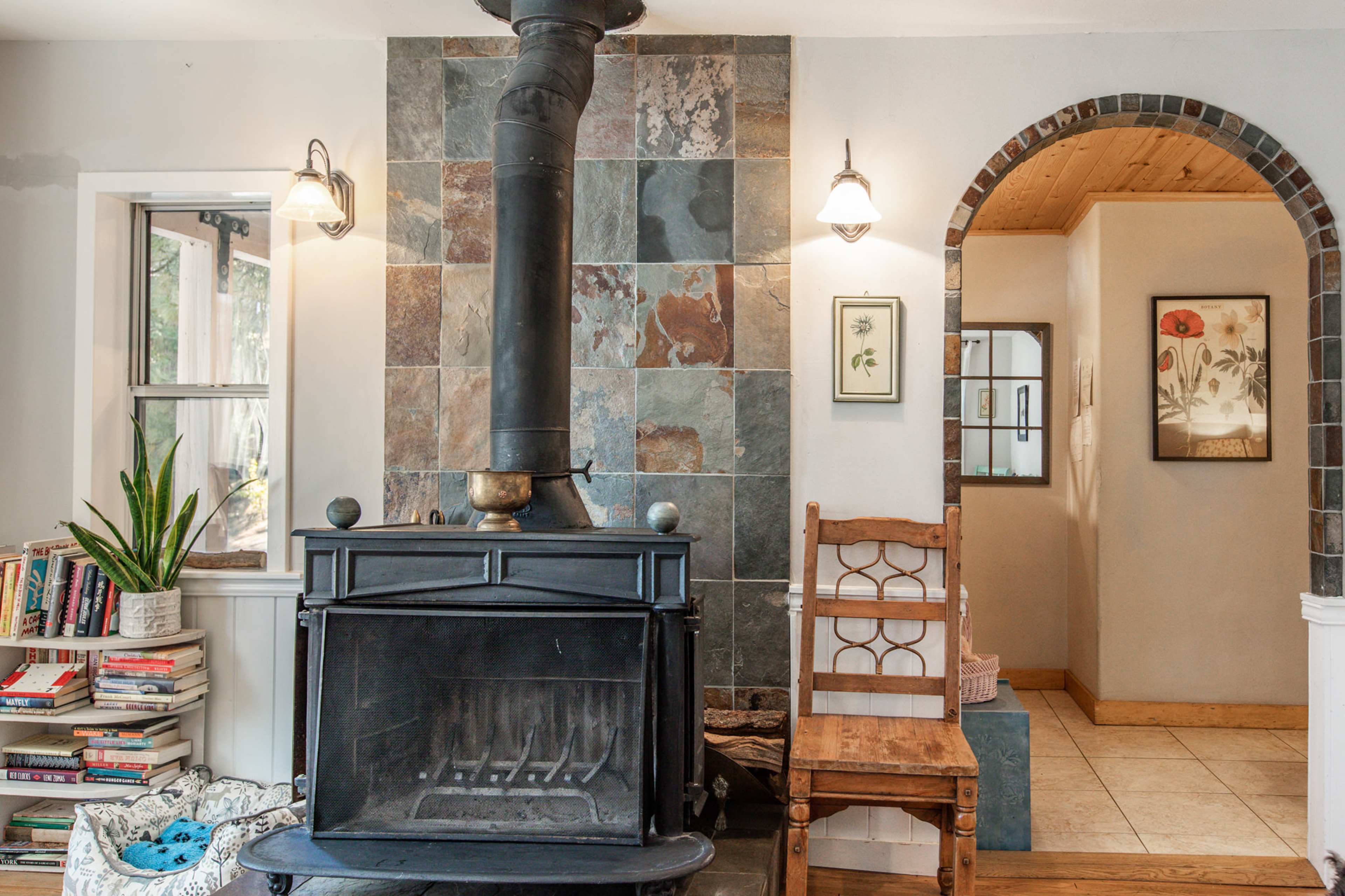 The image shows a living space with a black wood stove next to a tiled wall, surrounded by bookshelves, a wooden chair, and an arched doorway leading to another room.